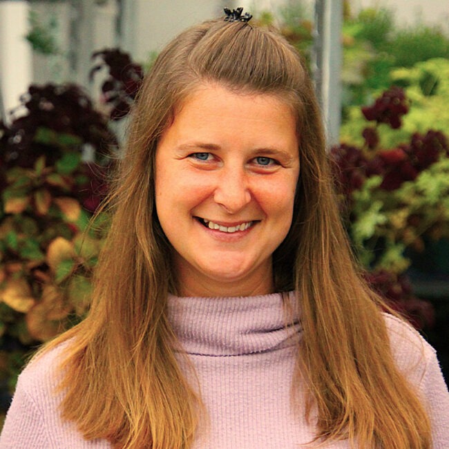 A person with long hair smiles at the camera, standing in front of greenery and plants, wearing a light sweater.