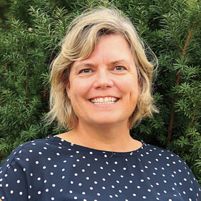 A smiling person with short hair wearing a polka-dotted shirt stands in front of green foliage, conveying a cheerful and relaxed atmosphere.