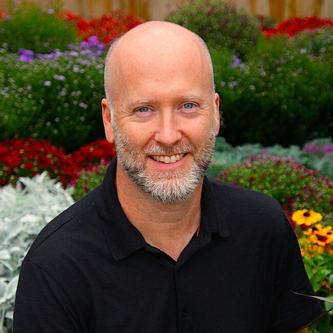 A person with a beard, wearing a black shirt, is smiling while sitting in a colorful garden filled with various flowers.