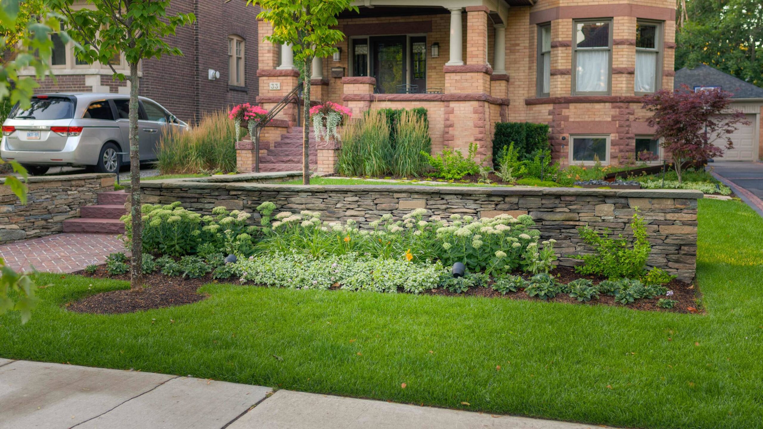 A brick house with stone landscaping, green lawn, flower beds, and a parked car on the driveway in a residential neighborhood.