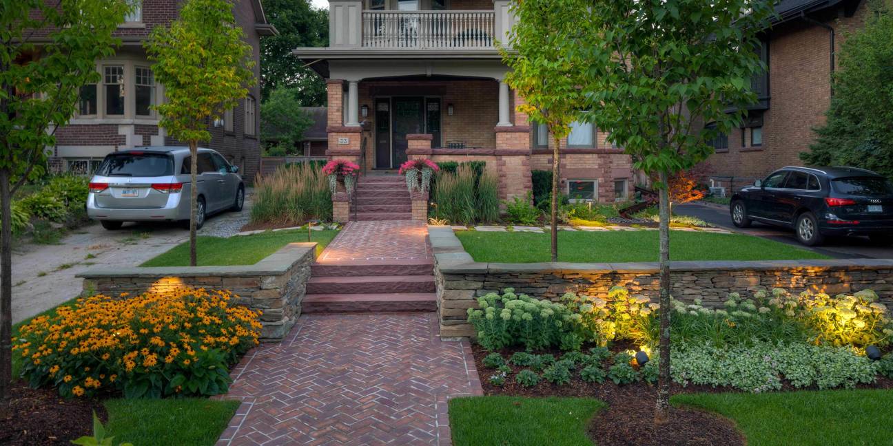 Brick house with manicured garden, two parked cars, and illuminated pathway. Abundant greenery and flowers create an inviting, picturesque residential scene.