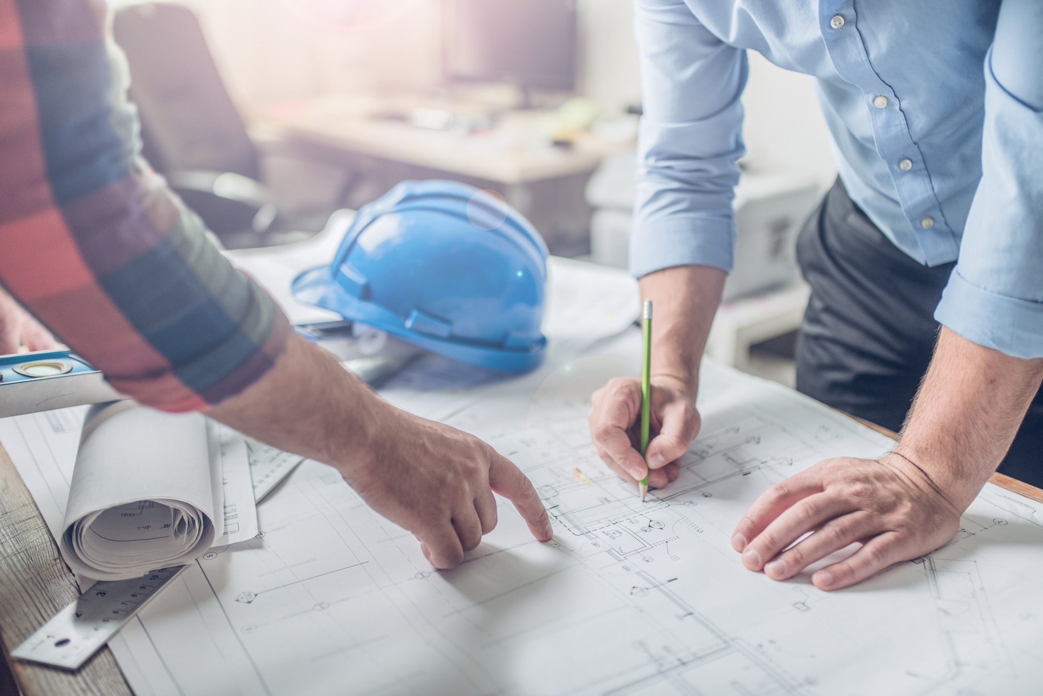Two people examining blueprints on a desk with a blue hard hat, pencil, and ruler, planning in a bright office setting.