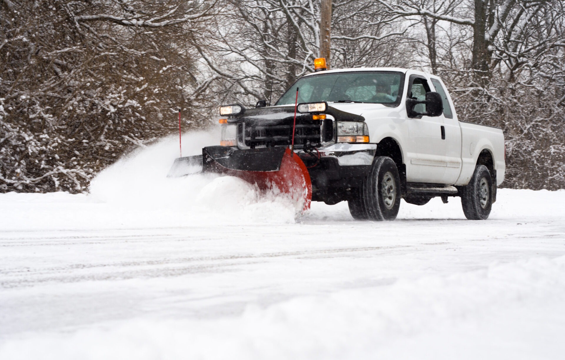 A snowplow truck clears snow on a road surrounded by bare trees, removing fresh snow from the ground, with no visible landmarks.