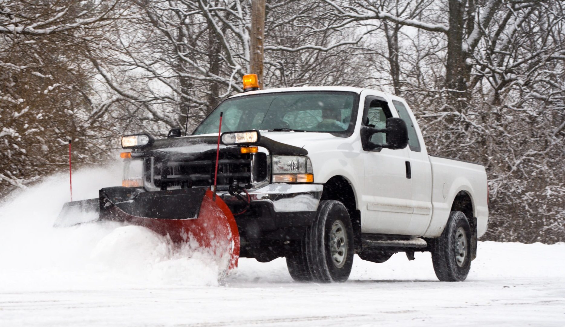 A snowplow truck clears snow on a road surrounded by bare trees, removing fresh snow from the ground, with no visible landmarks.
