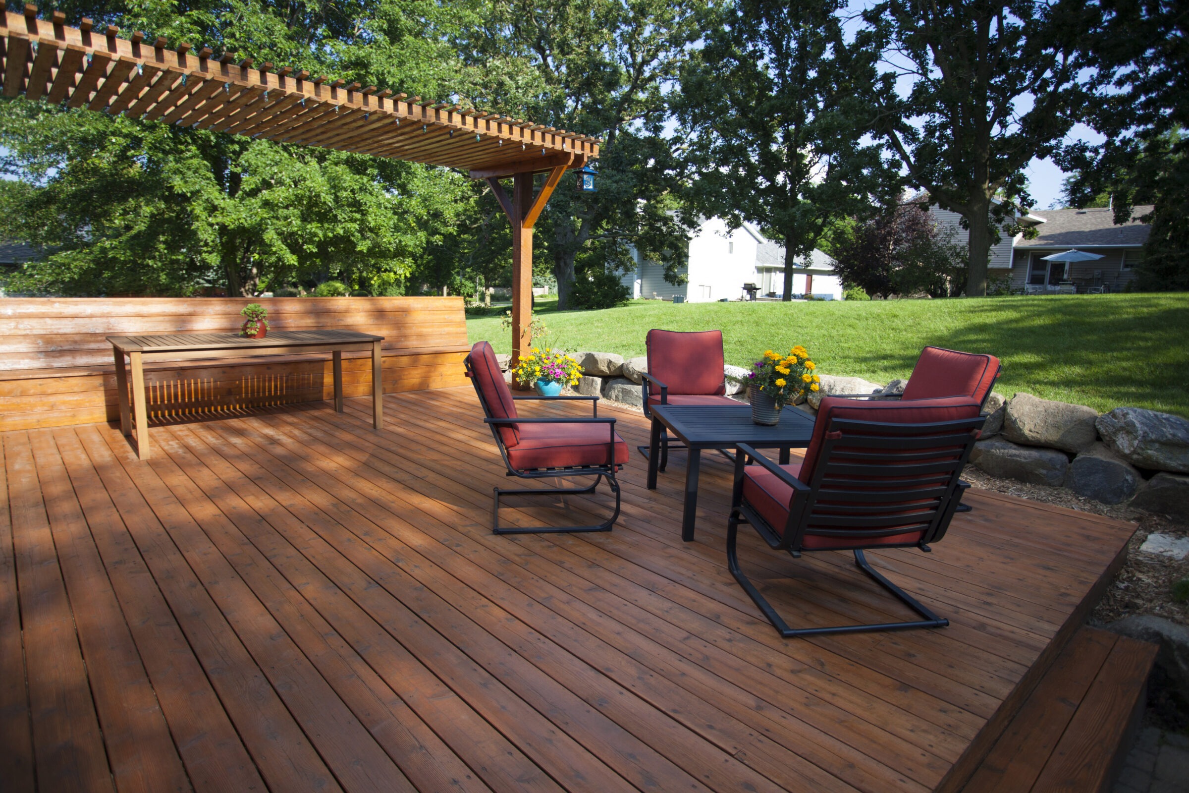 Wooden deck with pergola, red-cushioned chairs, and table. Surrounded by greenery and stones, creating a peaceful outdoor setting with colorful flowers.
