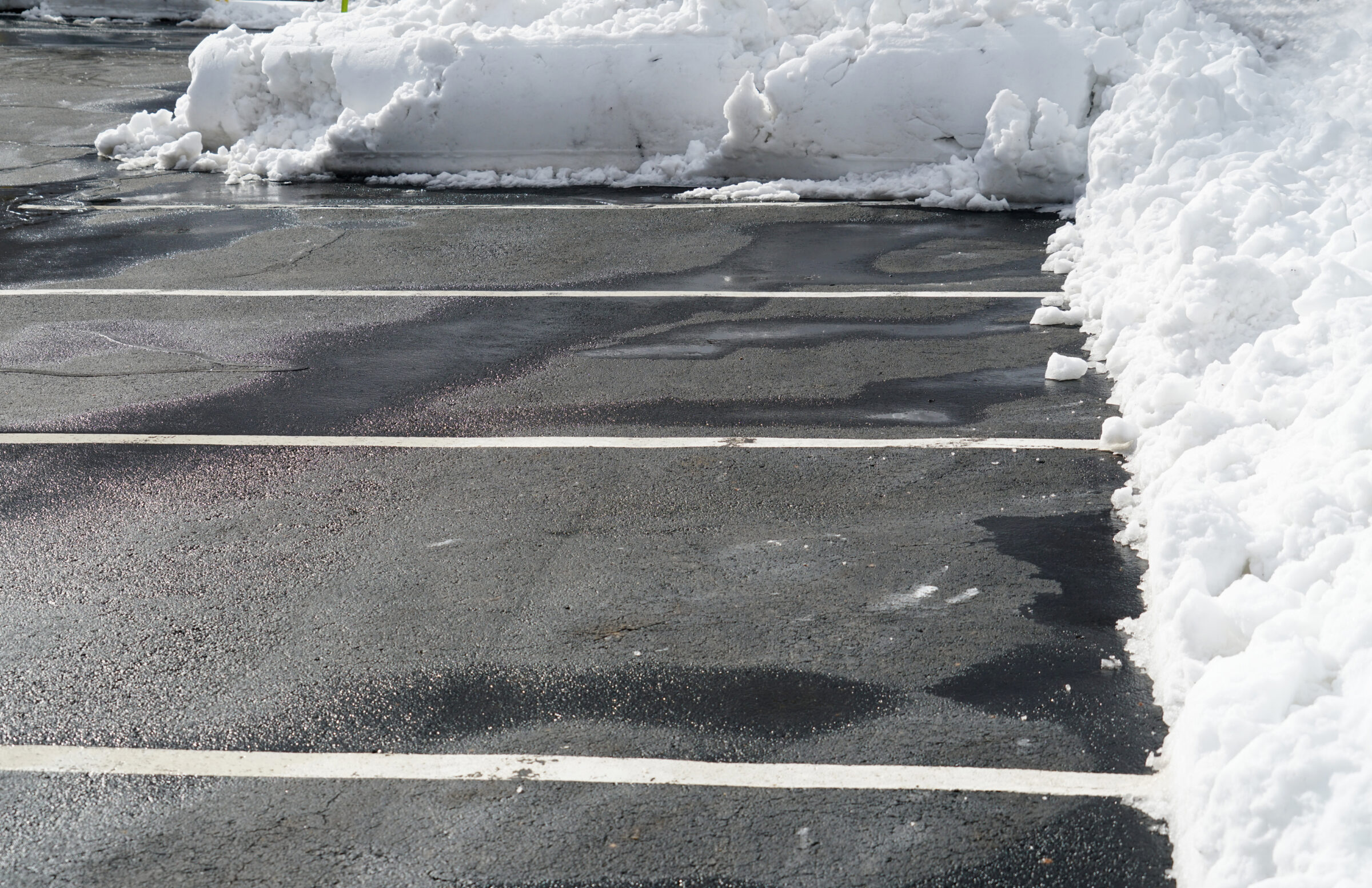 Snow-covered parking lot with empty spaces and piled snow around the edges, under a clear sky. No people or buildings visible.