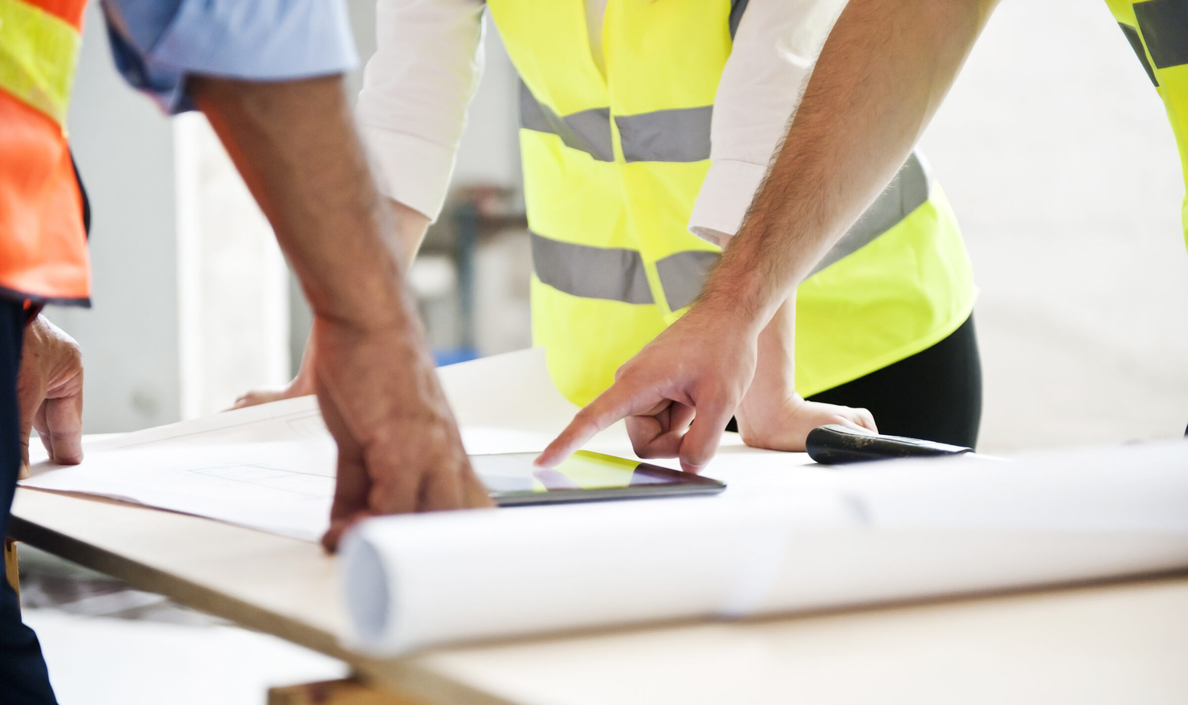 Two persons wearing safety vests examine blueprints on a table, pointing at a detail, in a collaborative construction environment.