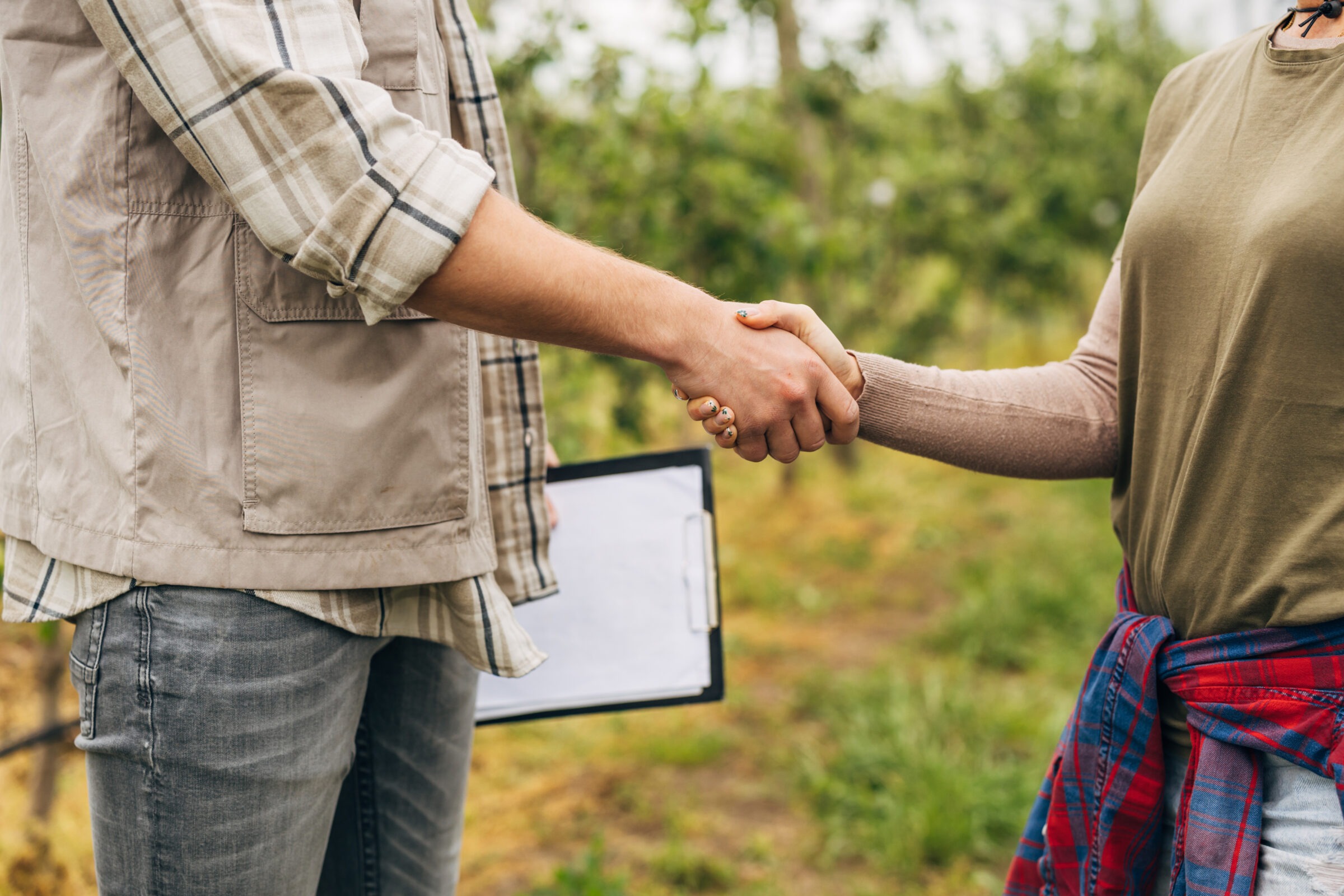 Two people shaking hands outdoors, one holds a clipboard, surrounded by greenery. The atmosphere is casual, with plaid and neutral clothing.