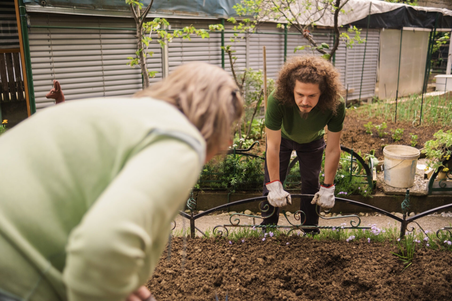 Two people gardening, engaging with soil and plants. Outdoor setting with greenery, garden tools, and protective gloves, focused on planting tasks.