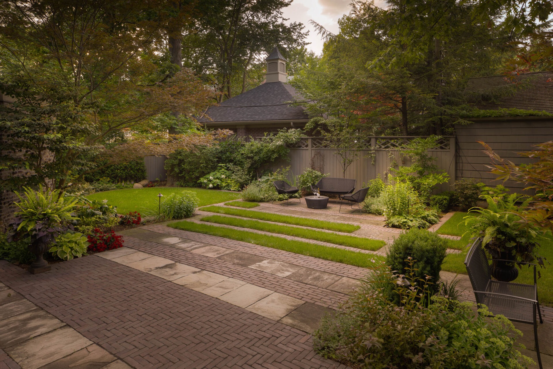 A serene, well-maintained garden with lush greenery, patio seating, stone pathways, and a small gazebo-like structure surrounded by tall trees.