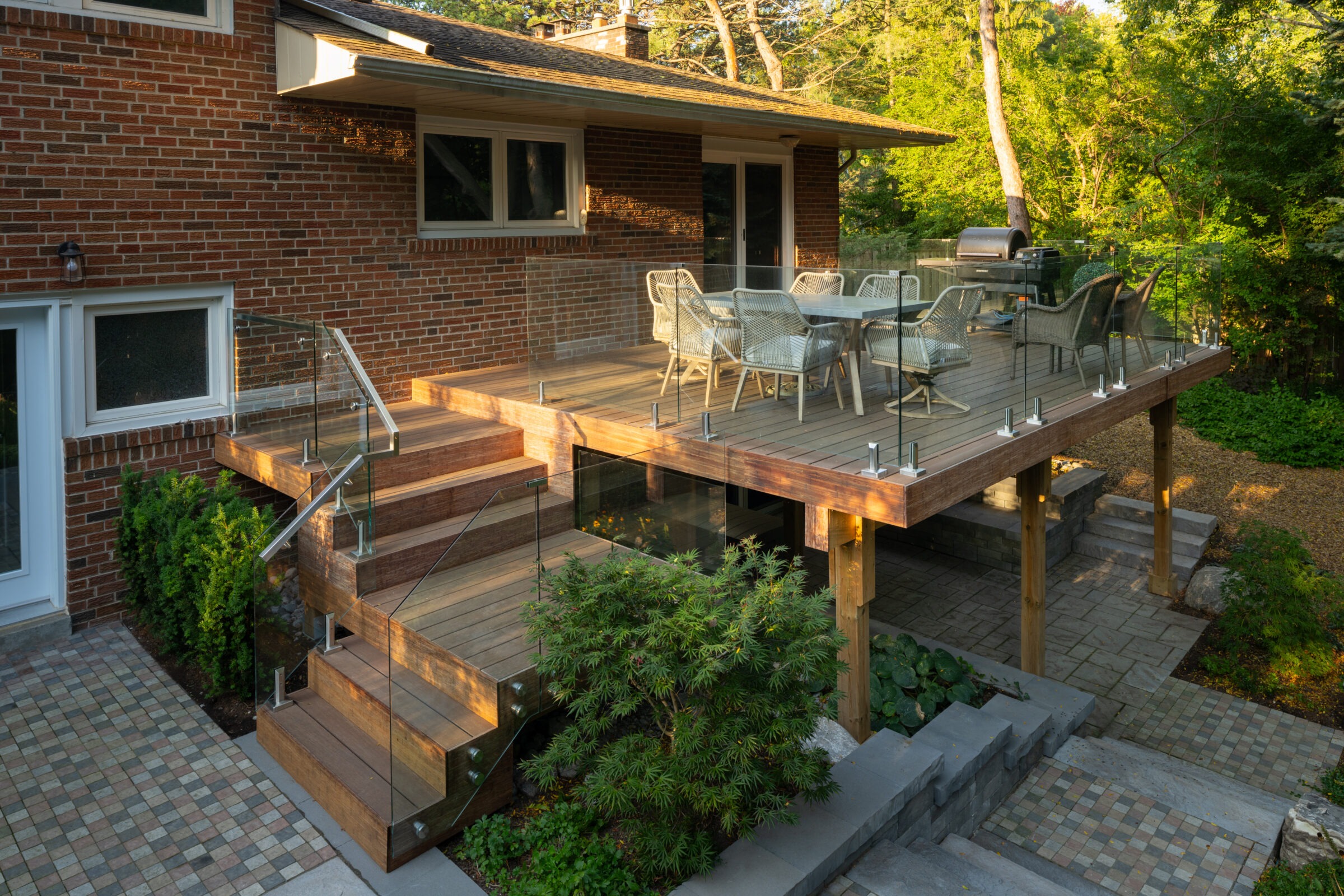 Modern elevated wooden deck attached to a brick house, featuring glass railings, a dining table with wicker chairs, a grill, and steps leading to a paved patio below, surrounded by landscaped greenery and trees.