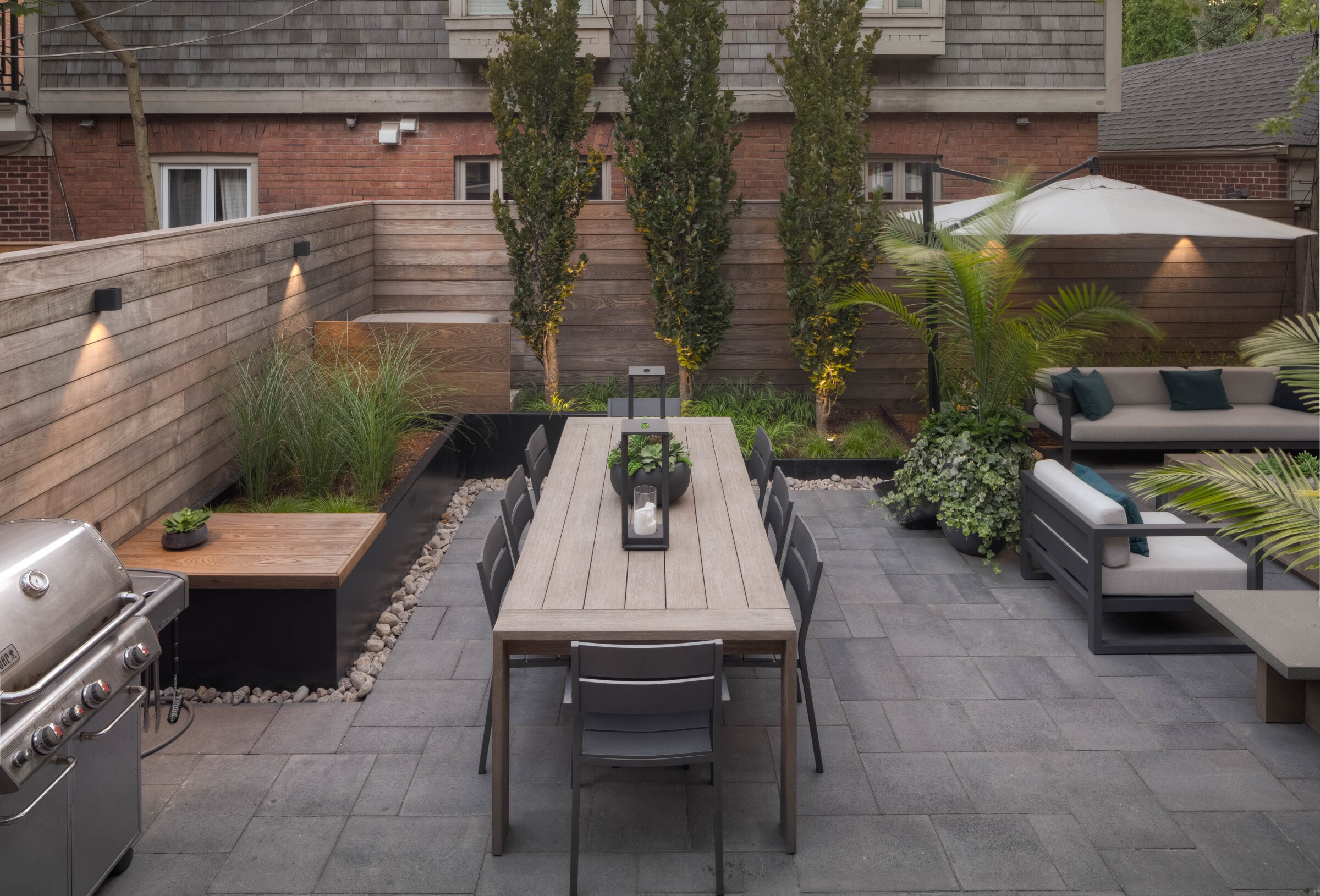 Modern backyard patio with a long wooden dining table, surrounding plants, gray stone flooring, a grill, and cozy seating under a large umbrella.