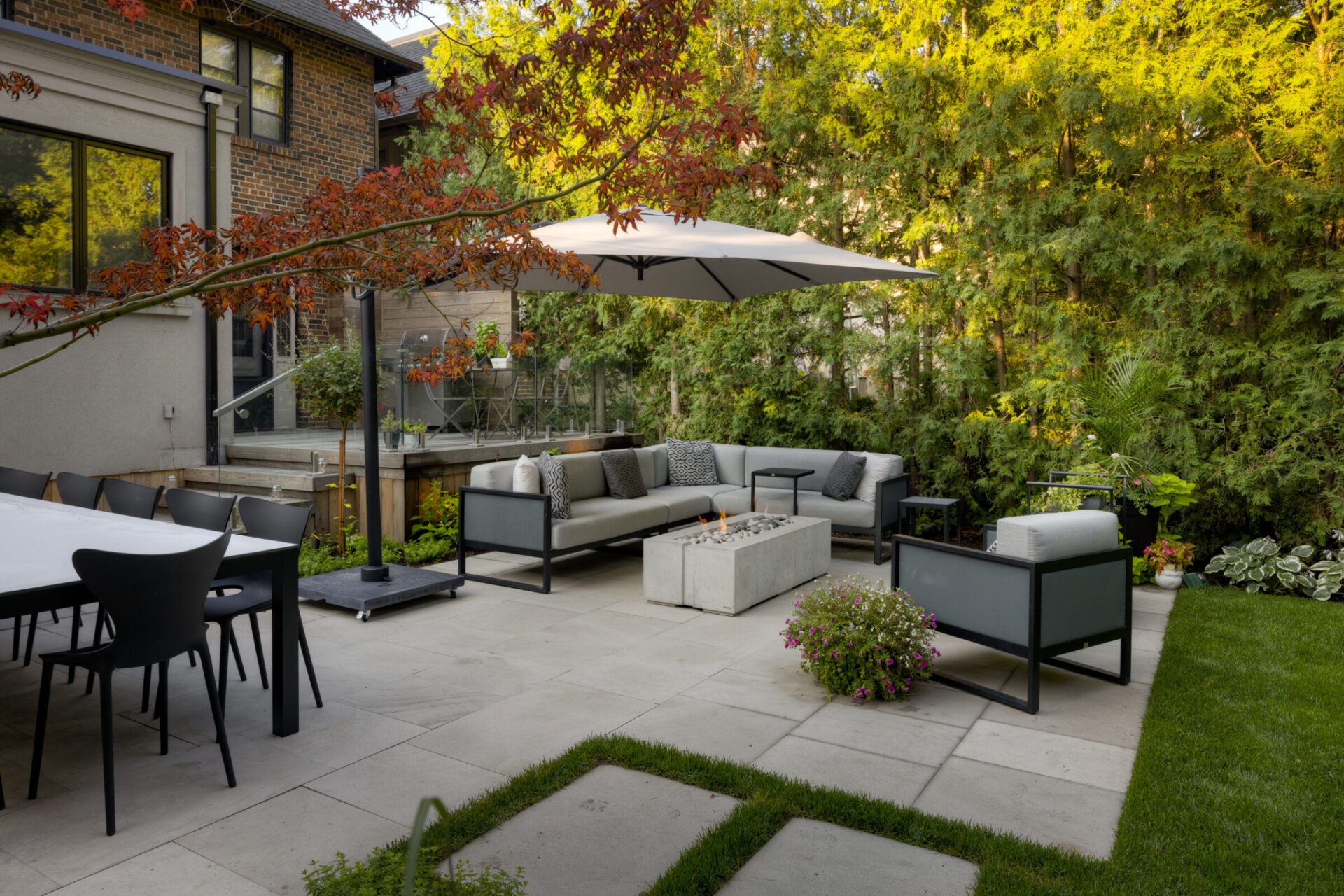 Modern backyard patio with gray sofa, fire pit, large umbrella, and table surrounded by chairs; lush greenery and brick house in background.