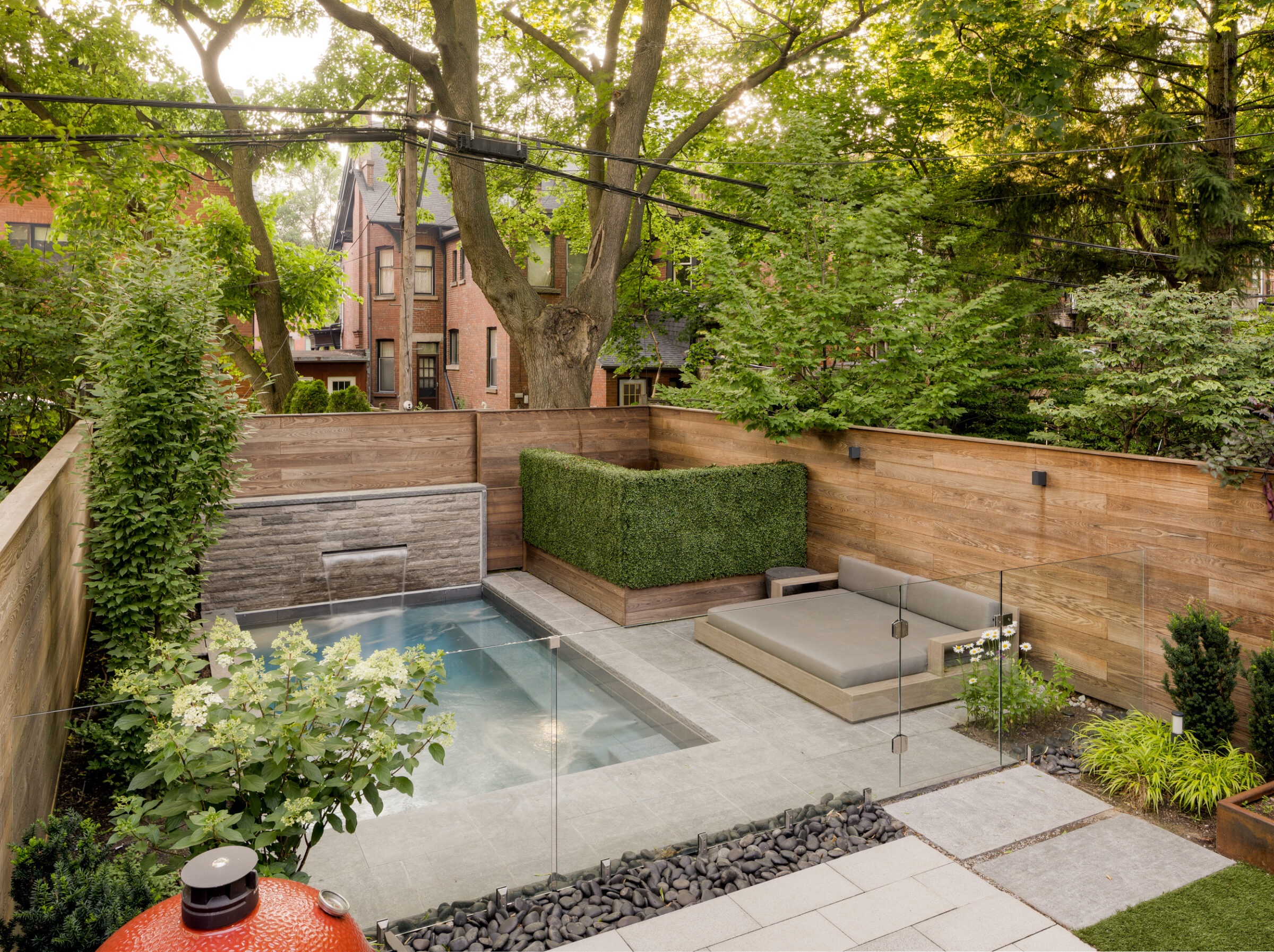 Modern backyard features a small pool, wooden fencing, and sleek seating area surrounded by lush greenery. Brick houses visible in the background.