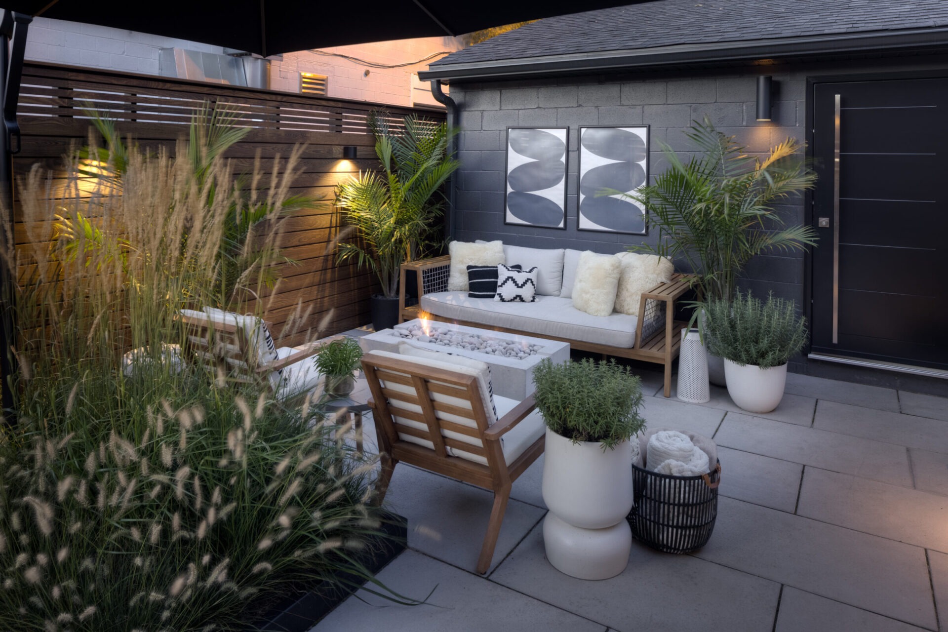 Modern patio with comfortable seating, potted plants, decorative pillows, and a fire pit. Wooden fencing and wall art create a cozy ambiance.