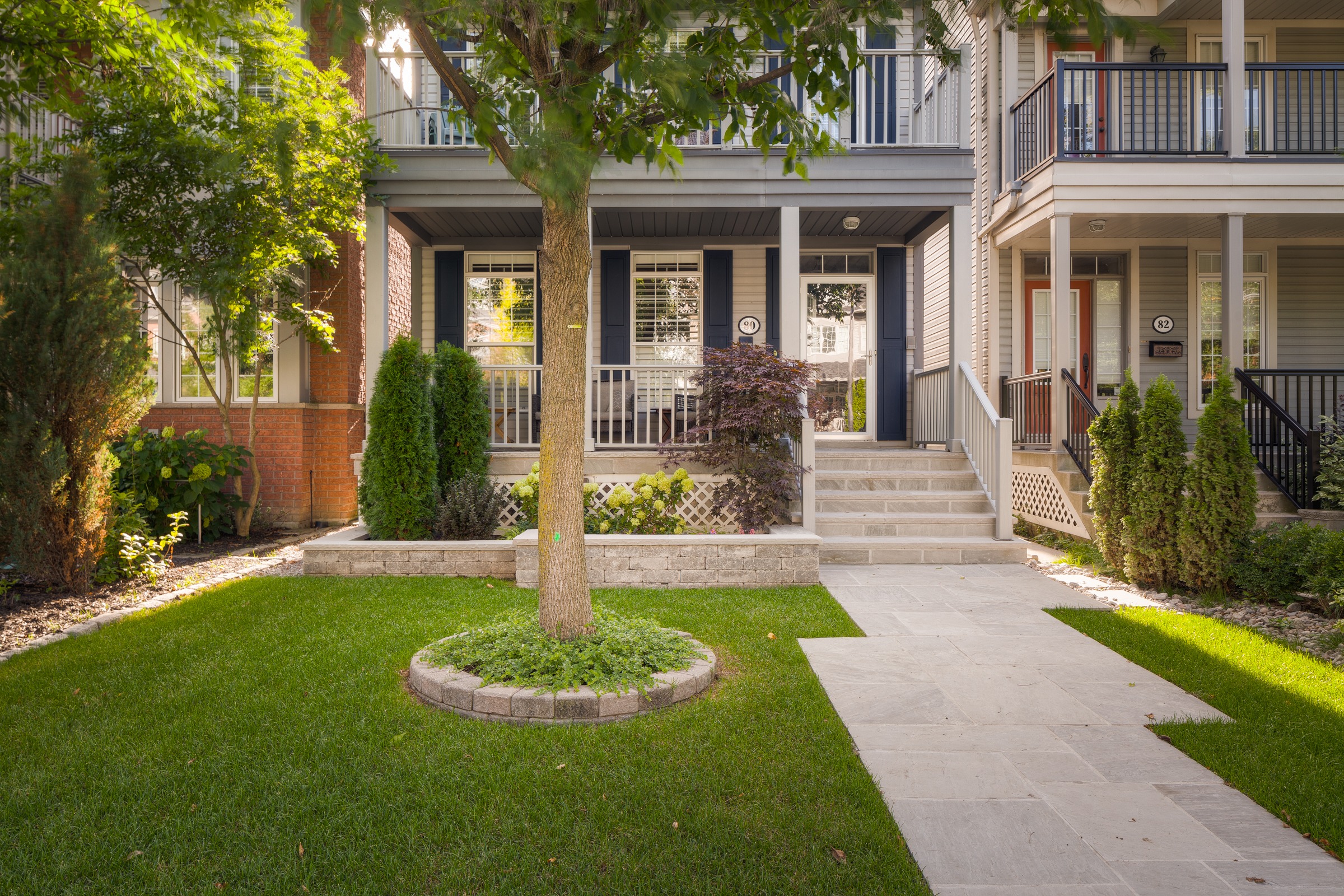 Charming townhouse-style front entrance with a tidy porch, manicured lawn, young tree surrounded by a stone ring, and a paved walkway leading to a set of stairs flanked by small shrubs and flower beds.