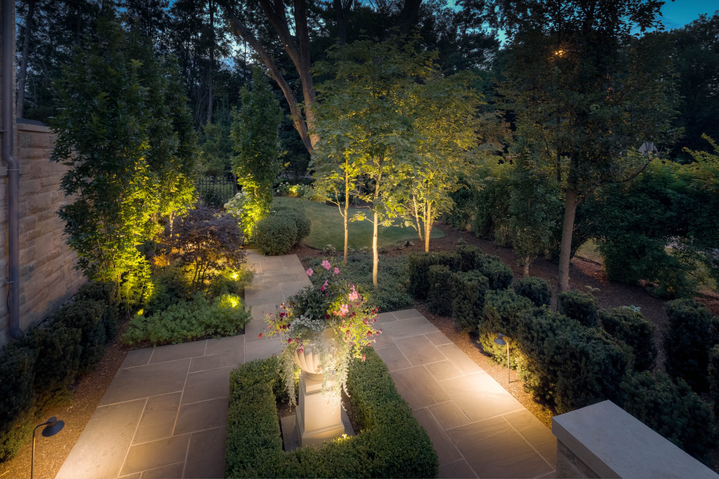 Upscale garden with a stone pathway, neatly trimmed hedges, and layered landscape lighting that highlights trees and flower beds. A central planter overflows with pink flowers in this serene dusk setting.
