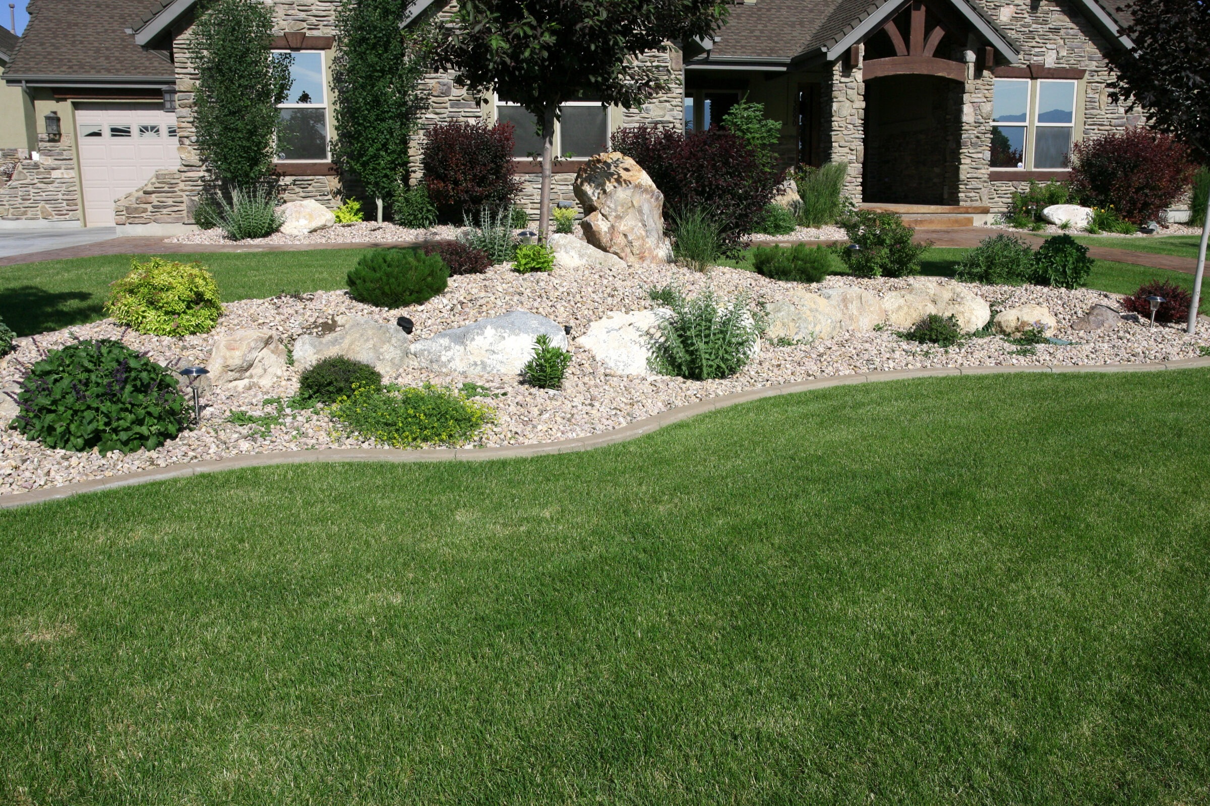 Xeriscaped front yard with drought-tolerant plants, large boulders, and decorative gravel, bordered by smooth concrete edging. Set in front of a stone house with a lush green lawn and manicured shrubs.