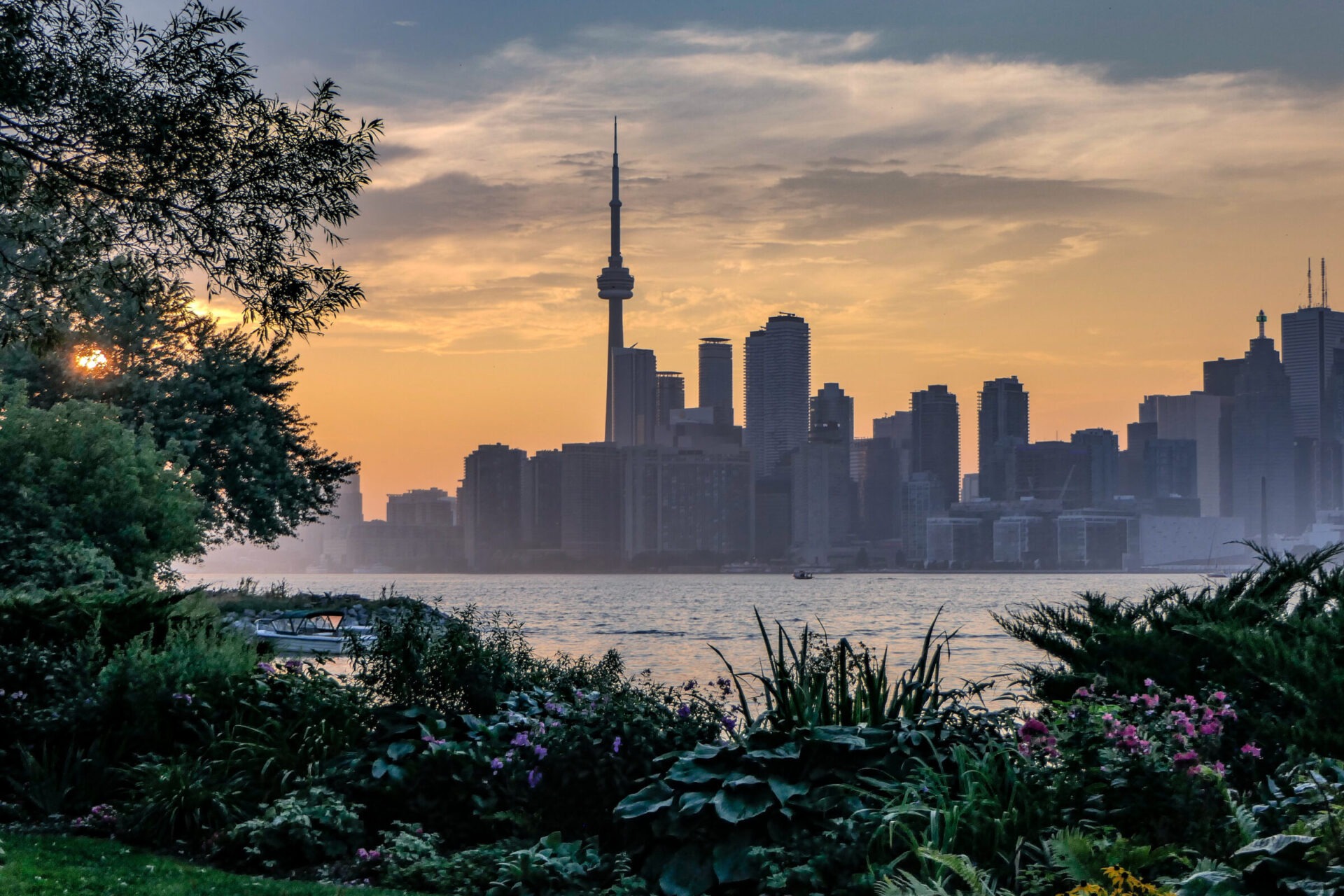 Toronto skyline at sunset with the CN Tower, seen from a lush garden across the water, creating a serene urban landscape.