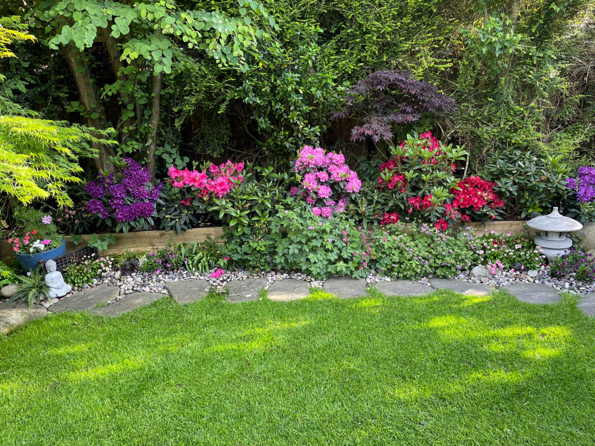 A vibrant garden features colorful flowers, lush greenery, a stone path, and a small Buddha statue, creating a serene, peaceful atmosphere.