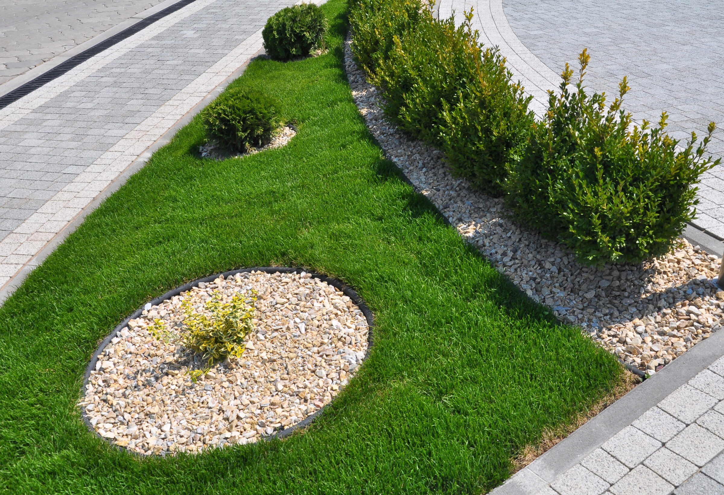 Modern low-maintenance landscape design with neatly trimmed grass, shrubs set in gravel mulch, and clean black metal edging. Surrounded by light gray paver walkways and driveway.