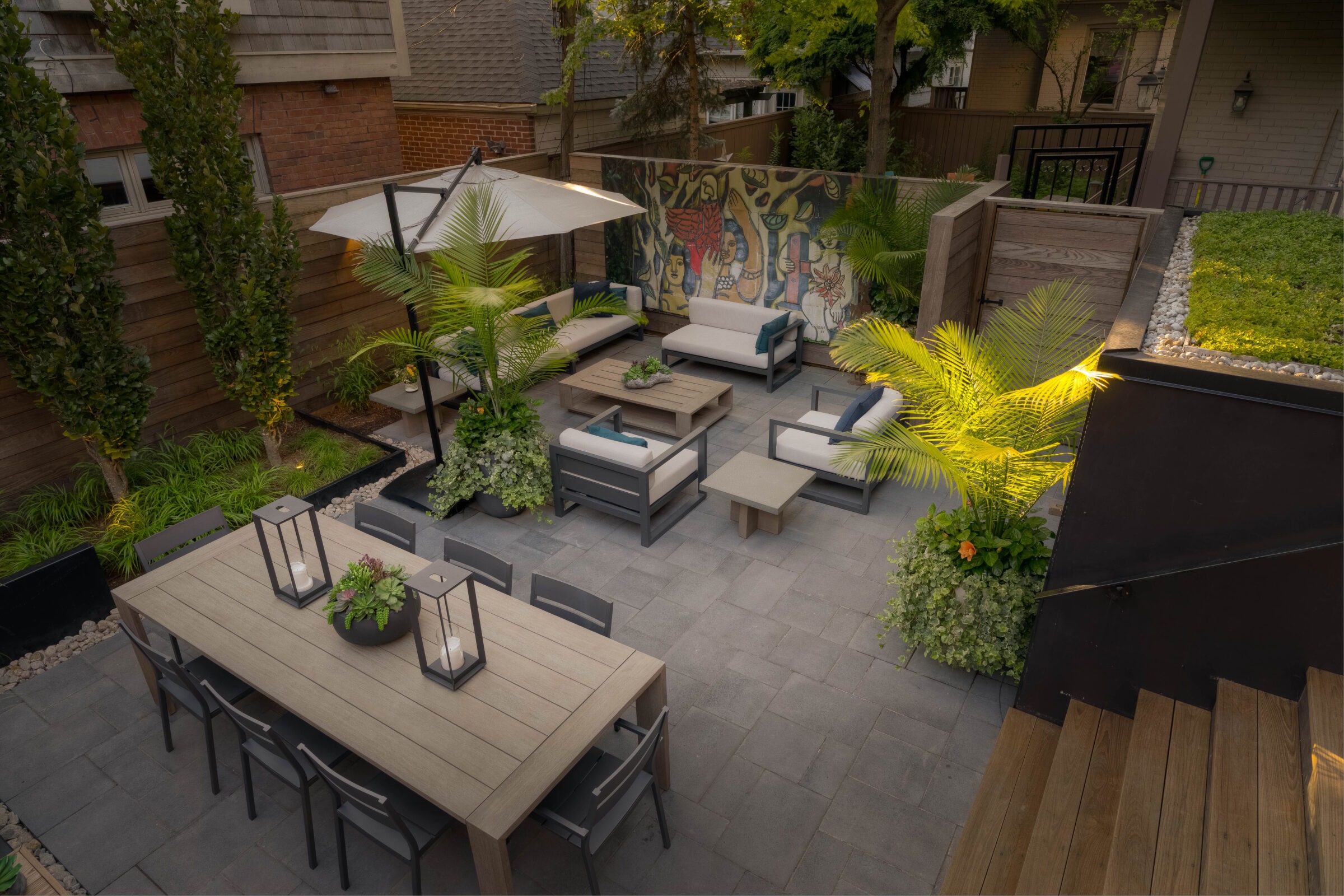 Stylish backyard patio featuring contemporary outdoor furniture, a dining table with lanterns, lush potted plants, and a colorful mural wall. The space is accented with soft lighting and surrounded by wood fencing and greenery.