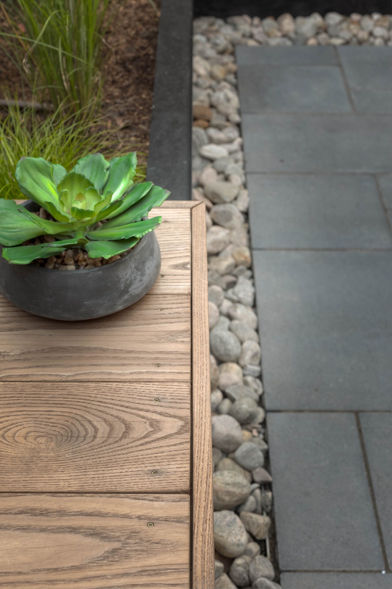 Close-up of a contemporary patio design featuring a wooden bench, gray paver stones, and a clean border of smooth river rocks. A potted succulent adds a touch of greenery to the minimalist setting.