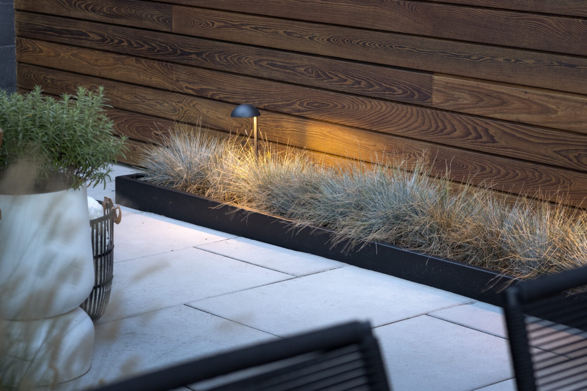 Sleek outdoor patio featuring a metal-edged planter with ornamental grasses, accented by a minimalist landscape light. Set against a wood-paneled wall and white paver flooring for a contemporary look.
