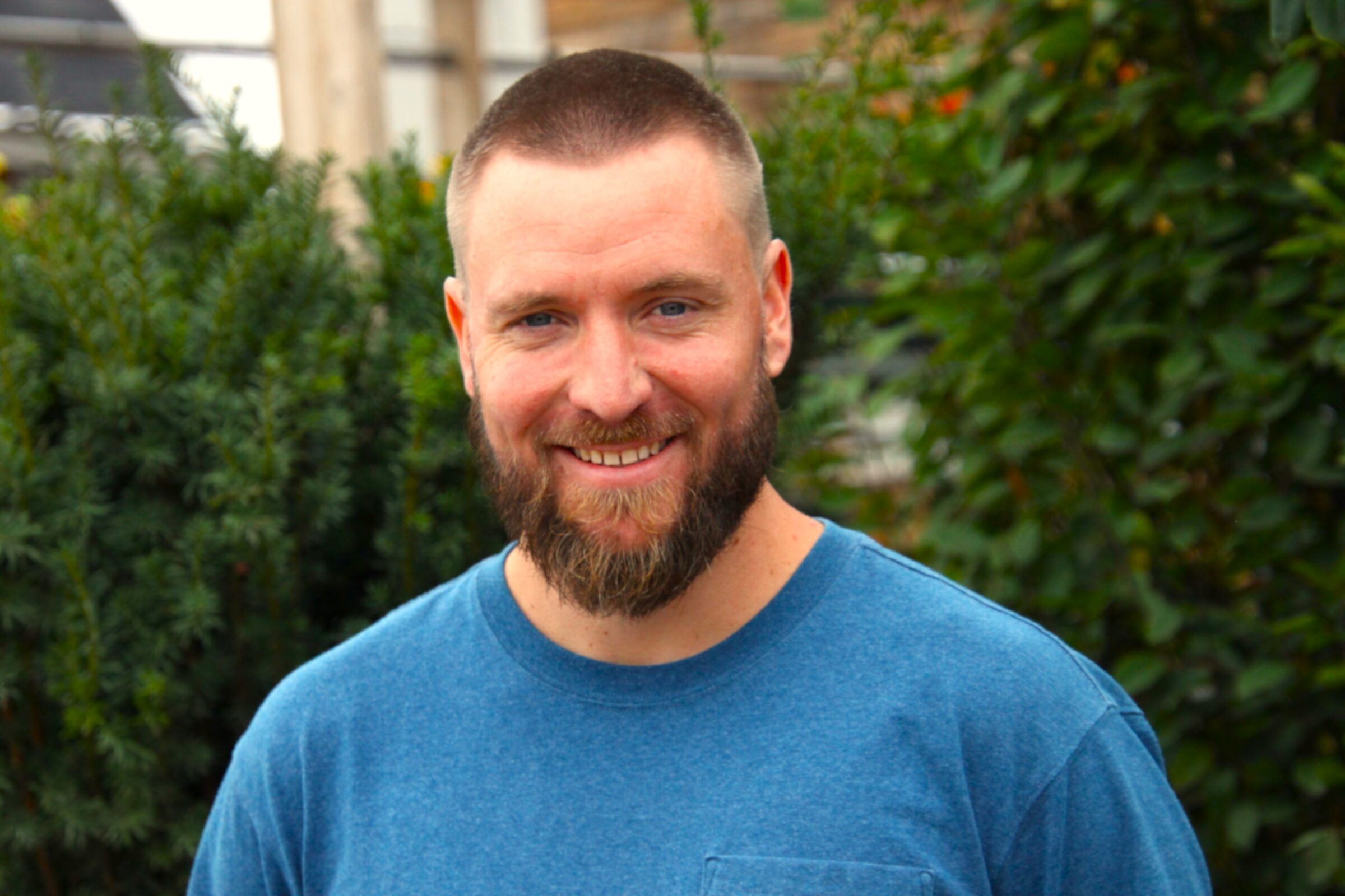 A person with a beard in a blue shirt smiles outdoors, standing in front of green shrubs and wooden structures in the background.