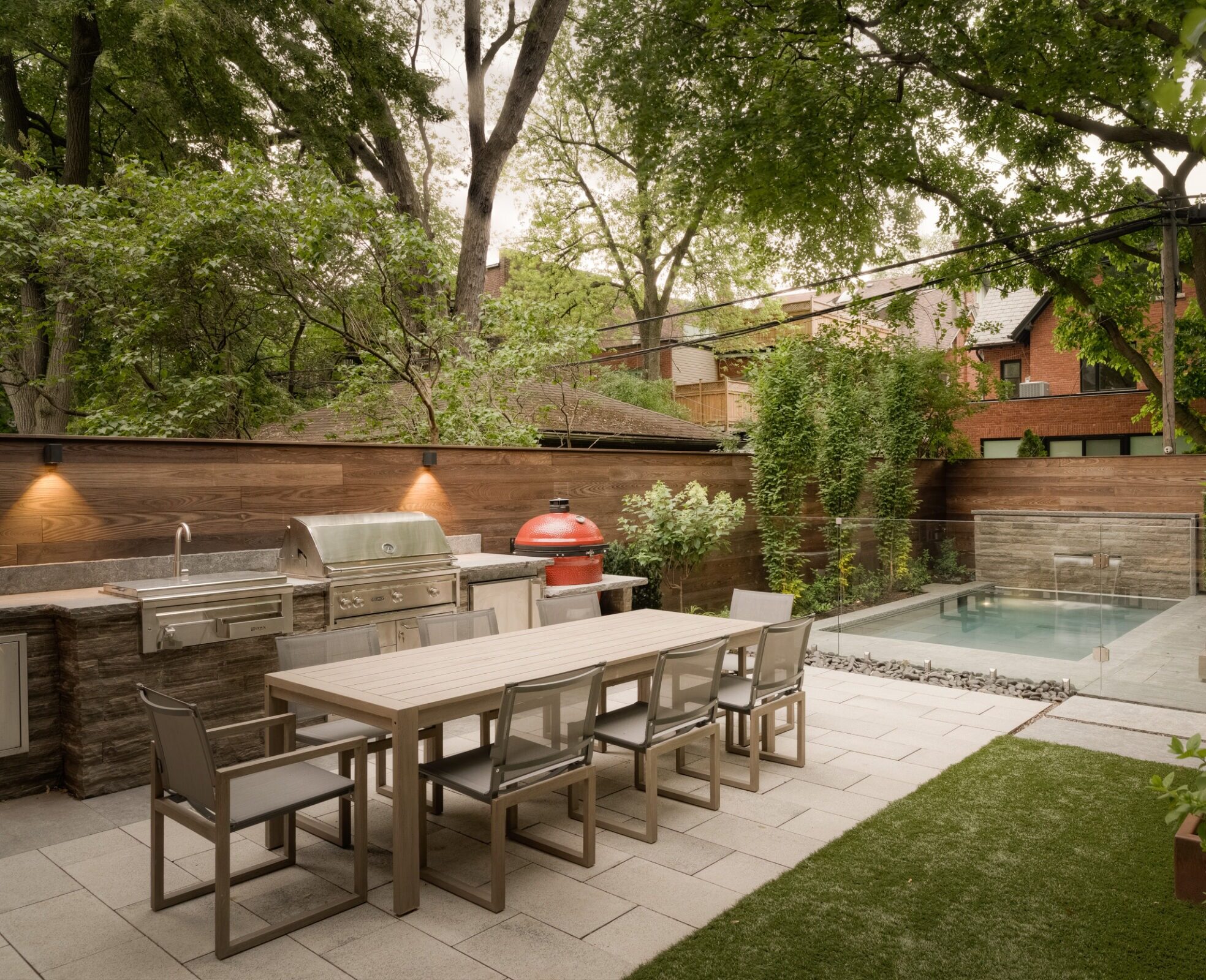 Modern backyard with dining table, grill, and small pool. Surrounded by lush trees and contemporary fencing. No recognizable landmarks or historical buildings visible.
