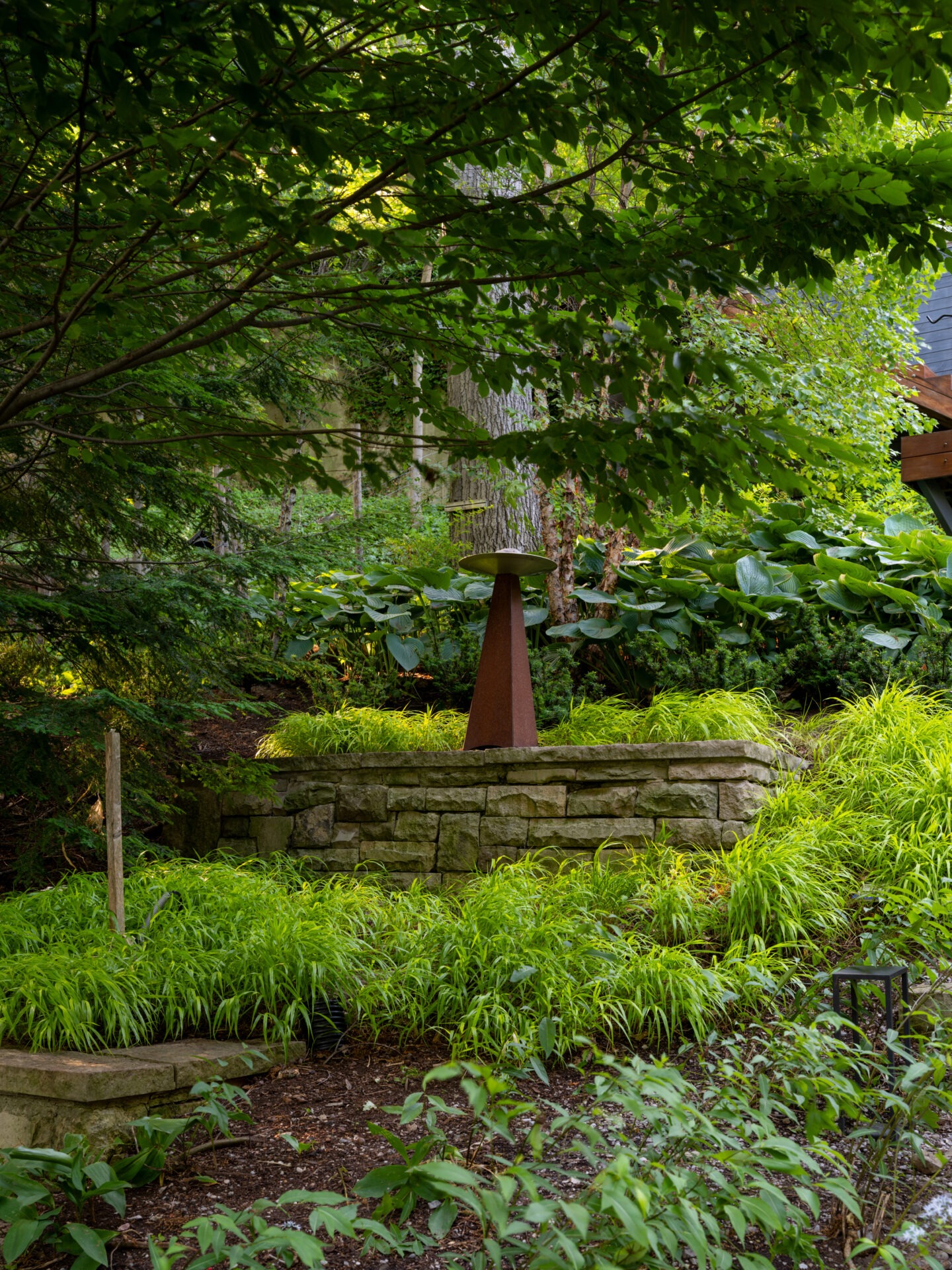 Lush green garden with a stone wall, prominent sculpture, and vibrant foliage under dappled sunlight. Tranquil, natural setting, surrounded by trees.