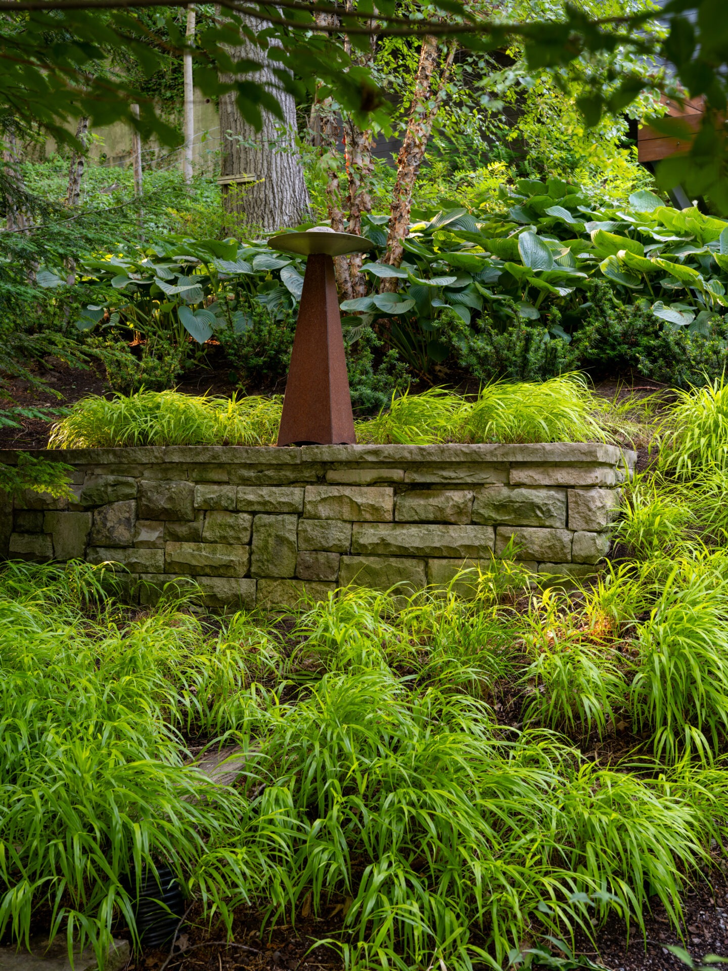 A rusted metal sculpture stands on a stone pedestal surrounded by lush, green plants and trees in a tranquil garden setting.