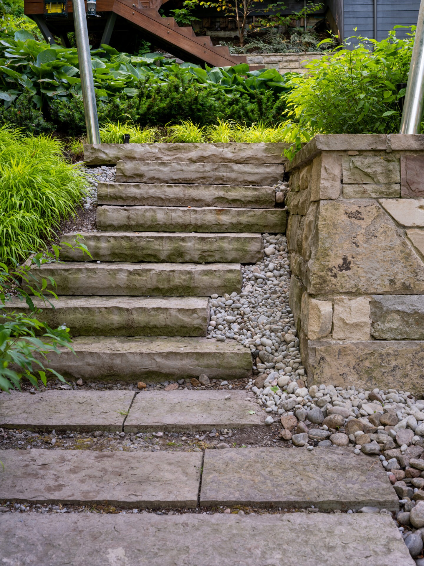 Stone steps and a gravel path are surrounded by lush, green plants. A wooden staircase is visible in the background.