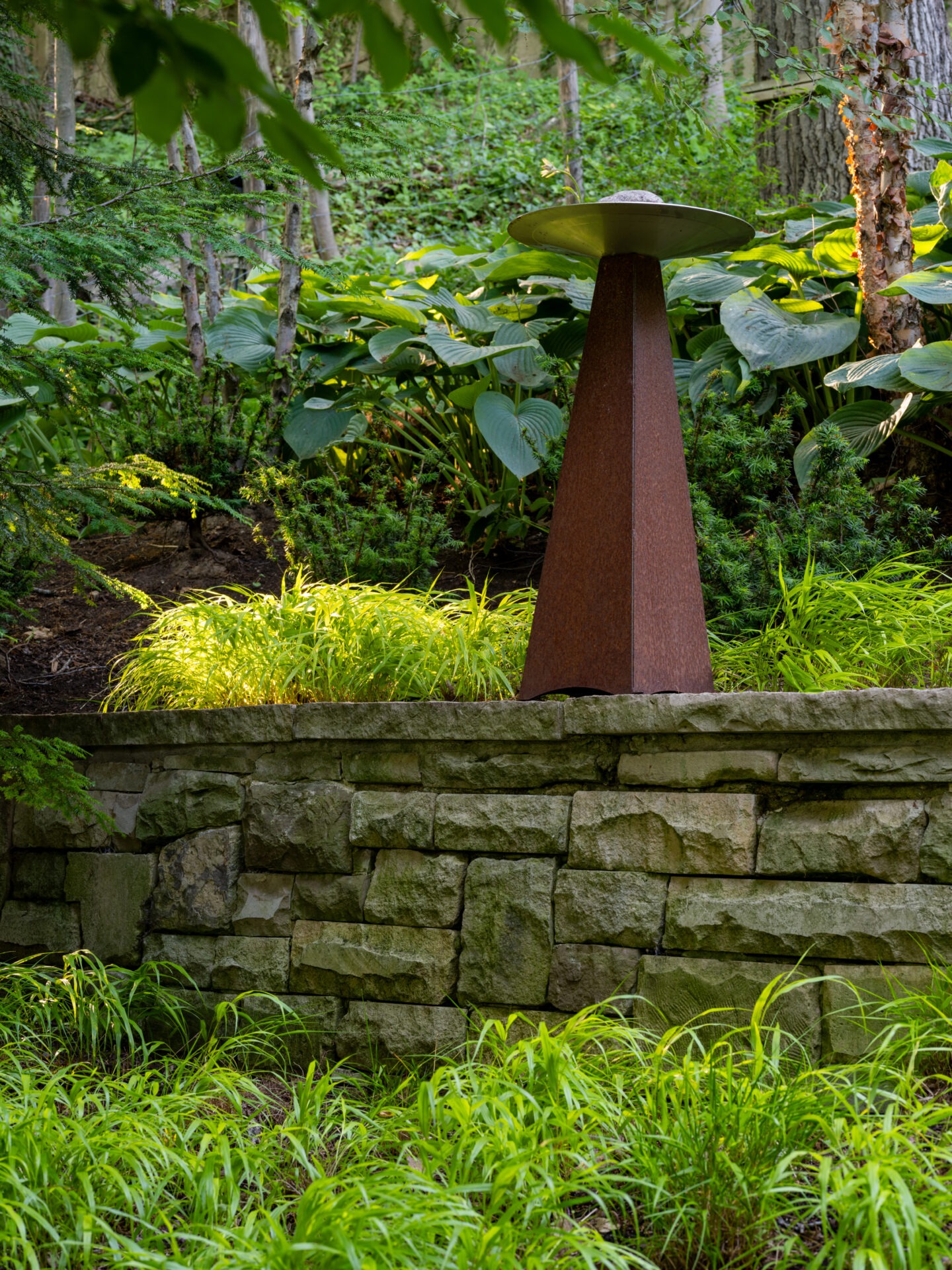 A rust-colored sculpture stands on a stone wall in a lush garden surrounded by trees and greenery, creating a tranquil, natural setting.