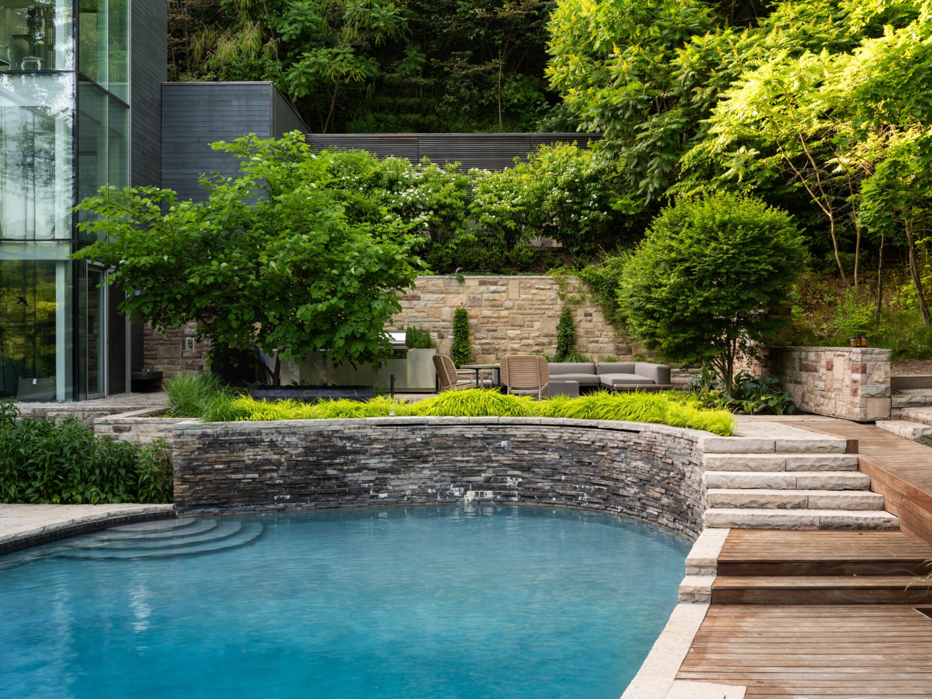Modern backyard with a glass-walled building, lush greenery, stone walls, a pool, wooden deck, and outdoor seating area amidst trees and bushes.