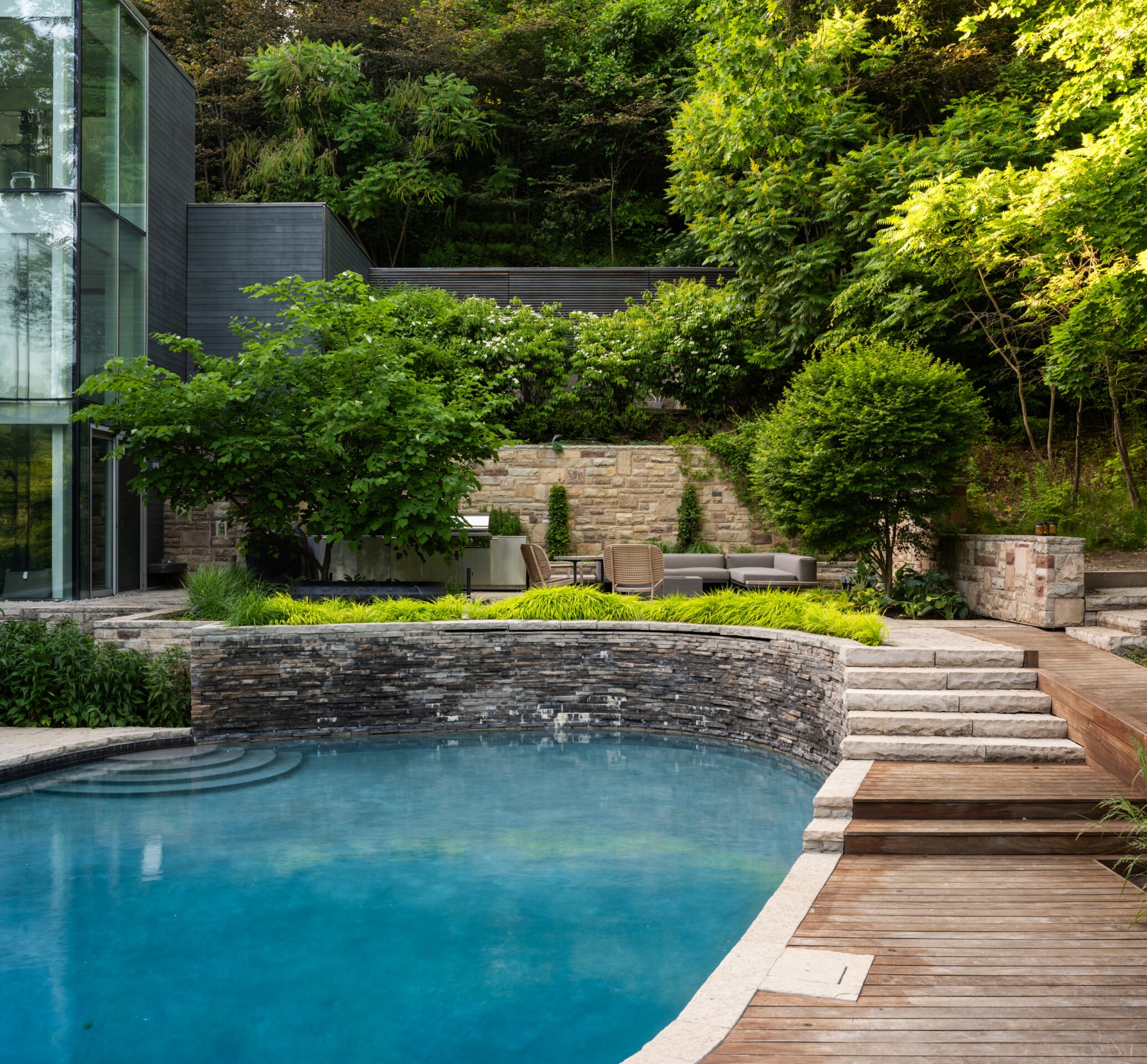 Modern glass building beside a serene pool surrounded by lush greenery and stone landscaping, creating a tranquil outdoor space.