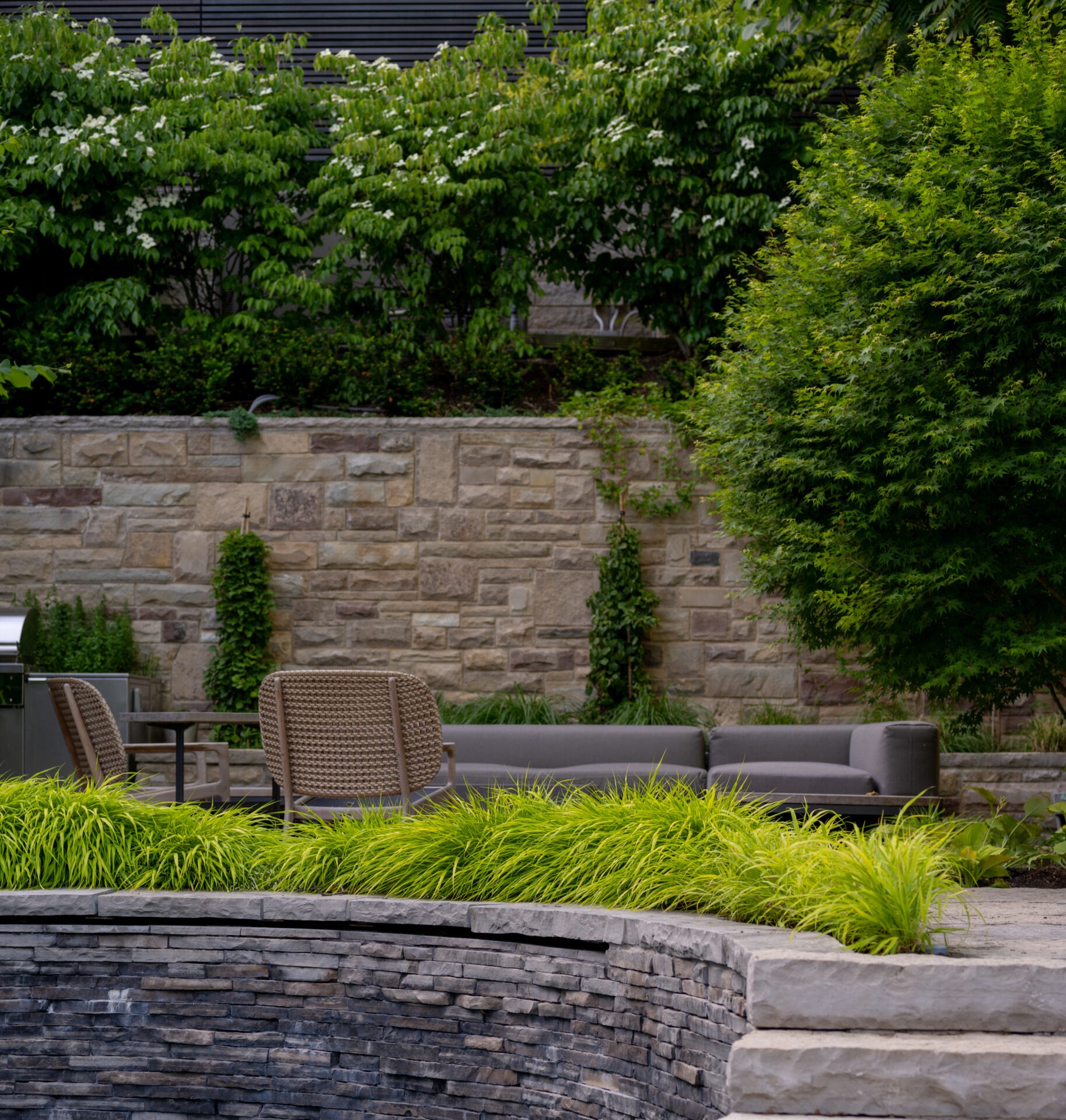 A peaceful garden patio with stone walls, lush greenery, outdoor seating, and a grill, perfect for relaxation and entertainment.