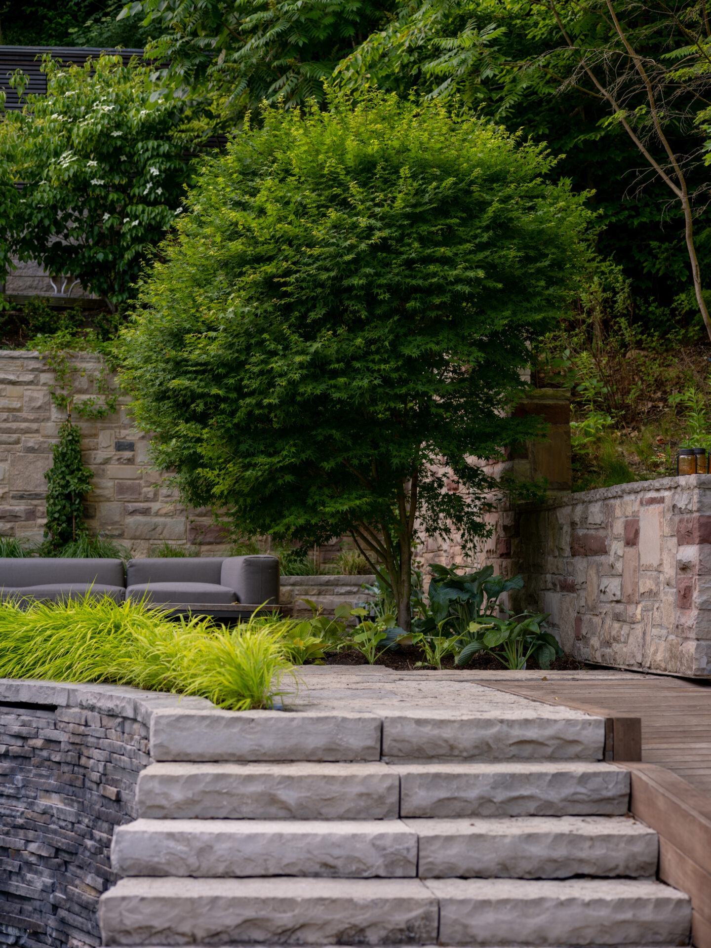 Stone steps lead to a serene garden terrace with a couch, surrounded by lush greenery and a stone wall backdrop.