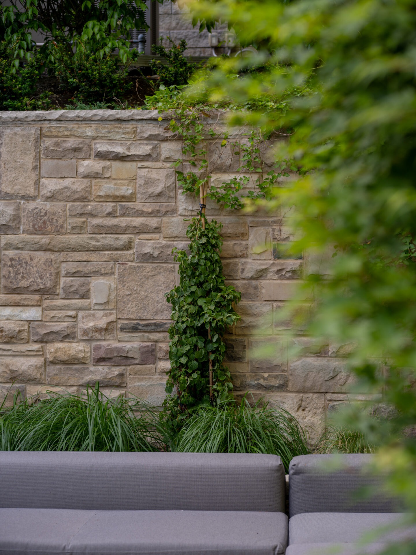 Gray seating with lush green plants in front of a rustic stone wall, creating a serene outdoor space with natural elements.
