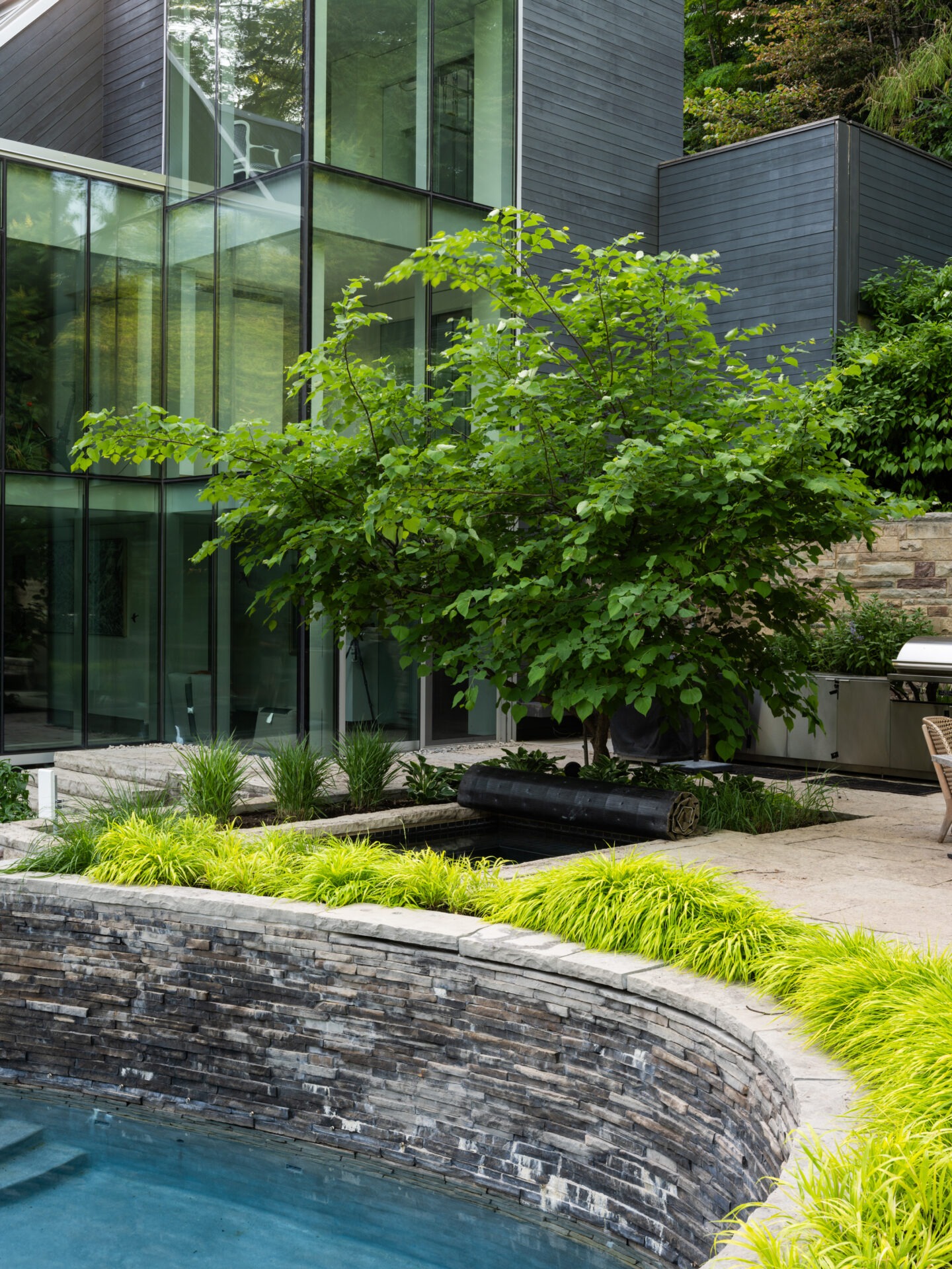 Modern house with glass exterior, lush greenery, and a stone-lined pool. The contemporary design integrates nature and architecture seamlessly, creating a serene atmosphere.