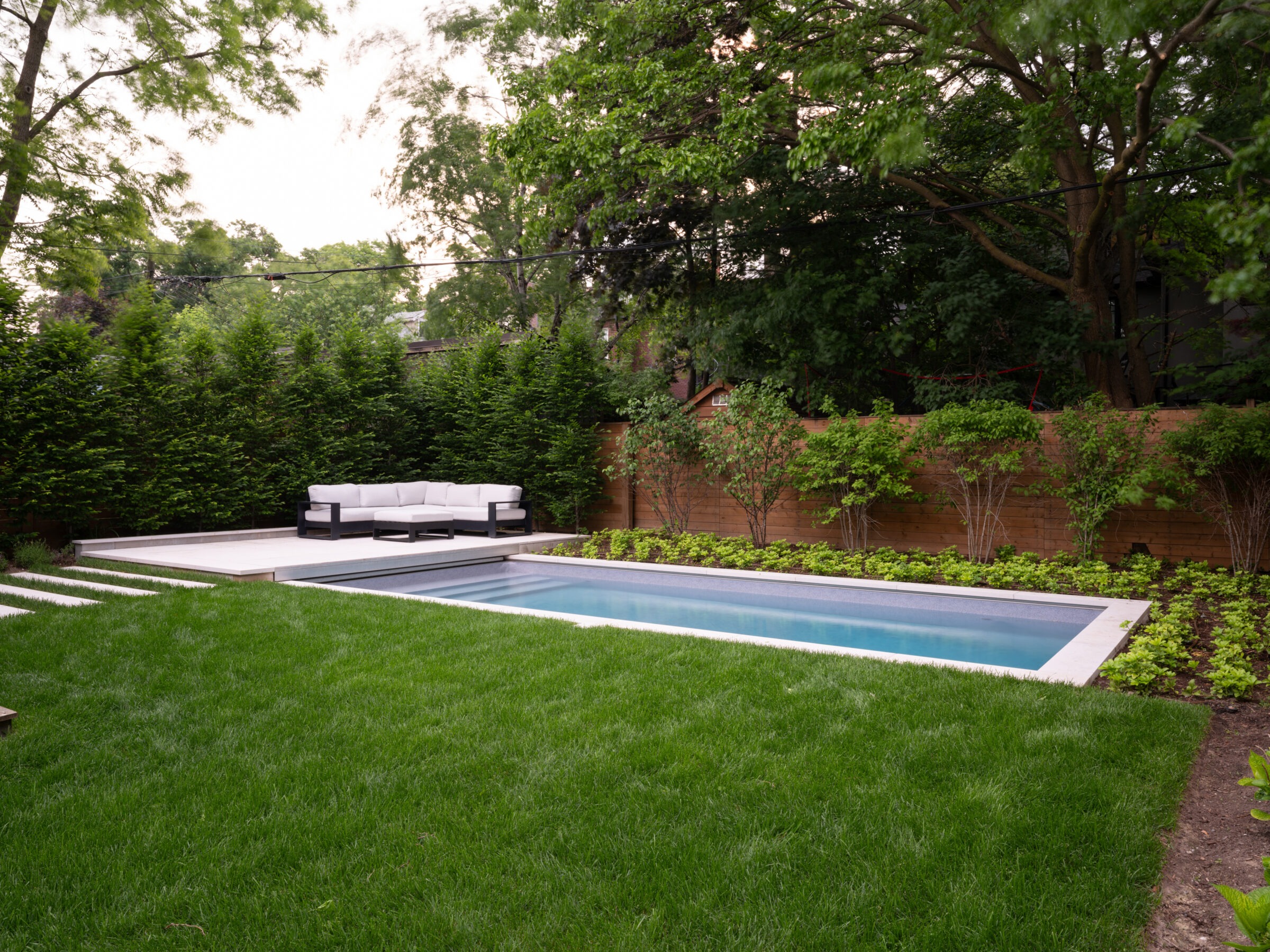 A serene backyard with a rectangular pool, surrounding greenery, and a comfortable white outdoor sofa set on a wooden deck.