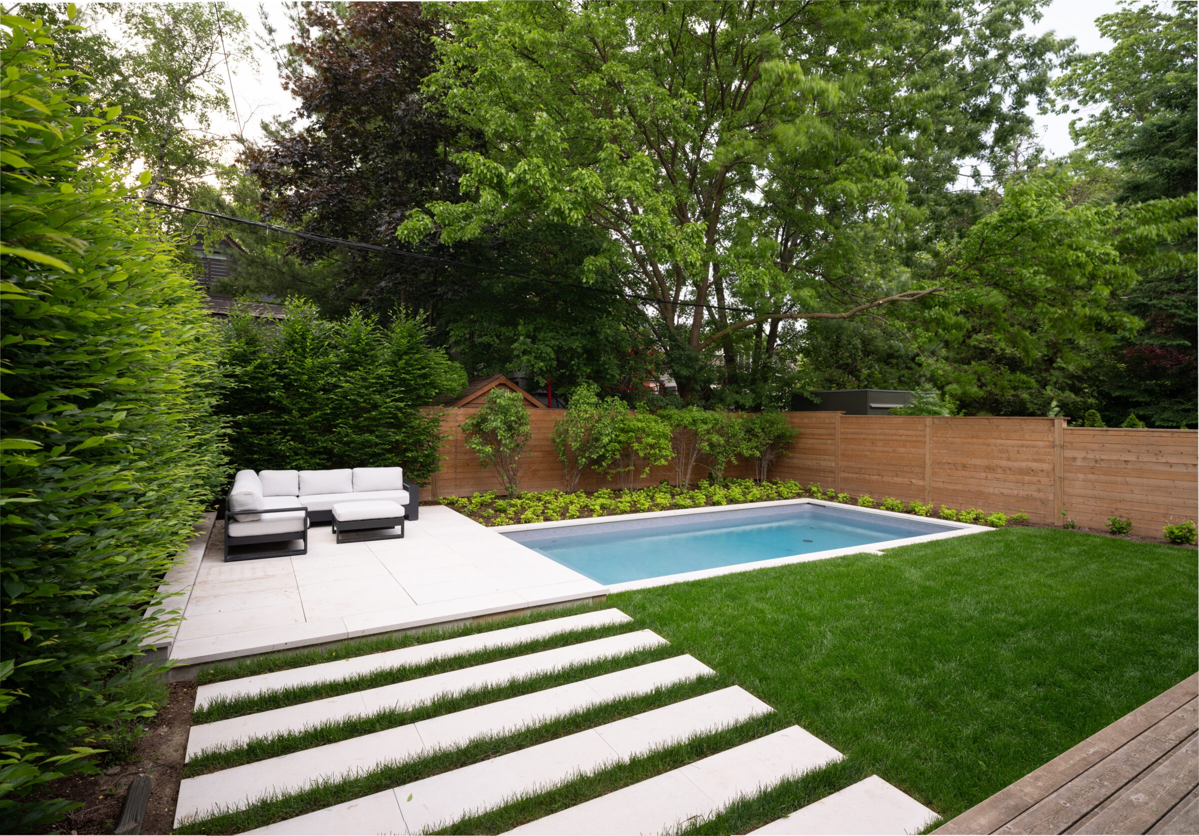 A modern backyard features a small pool, white seating area, and lush greenery, enclosed by a wooden fence and surrounded by trees.