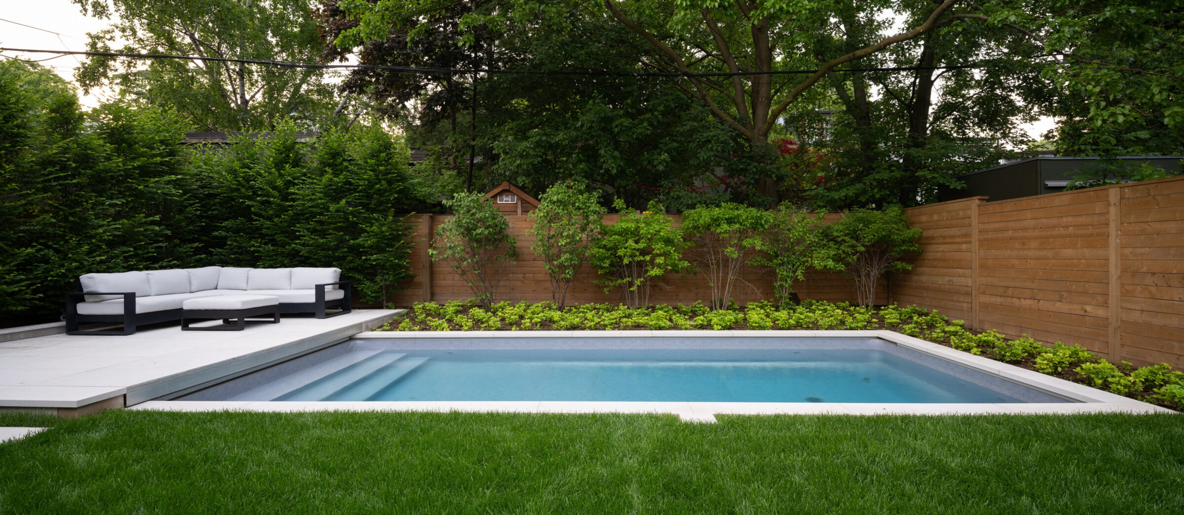 A backyard features a rectangular pool, surrounded by lush greenery, wooden fence, and grey patio with white outdoor sofa set.