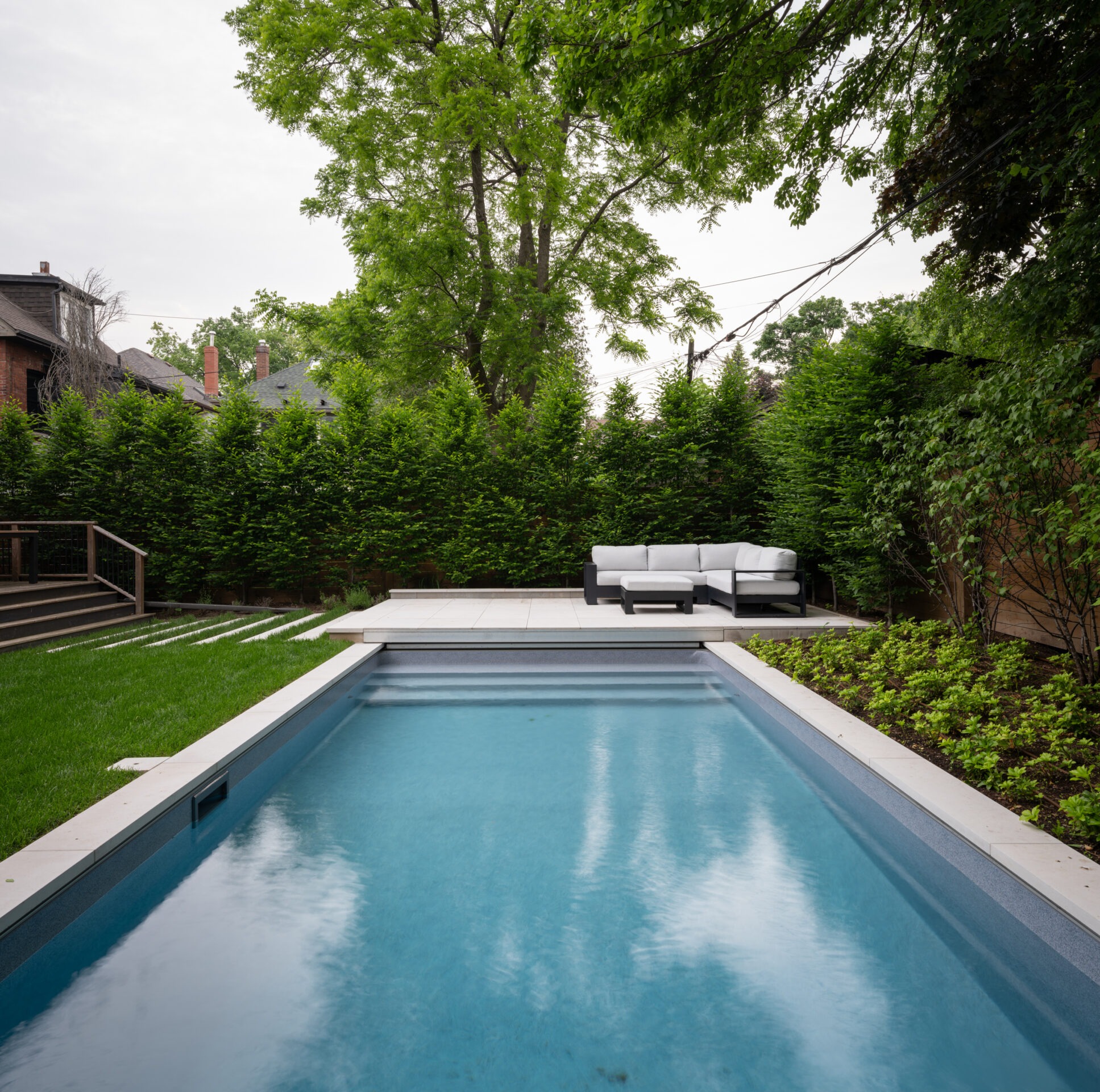 A modern backyard features a rectangular pool, lush greenery, a patio with white seating, and surrounding trees, creating a tranquil atmosphere.