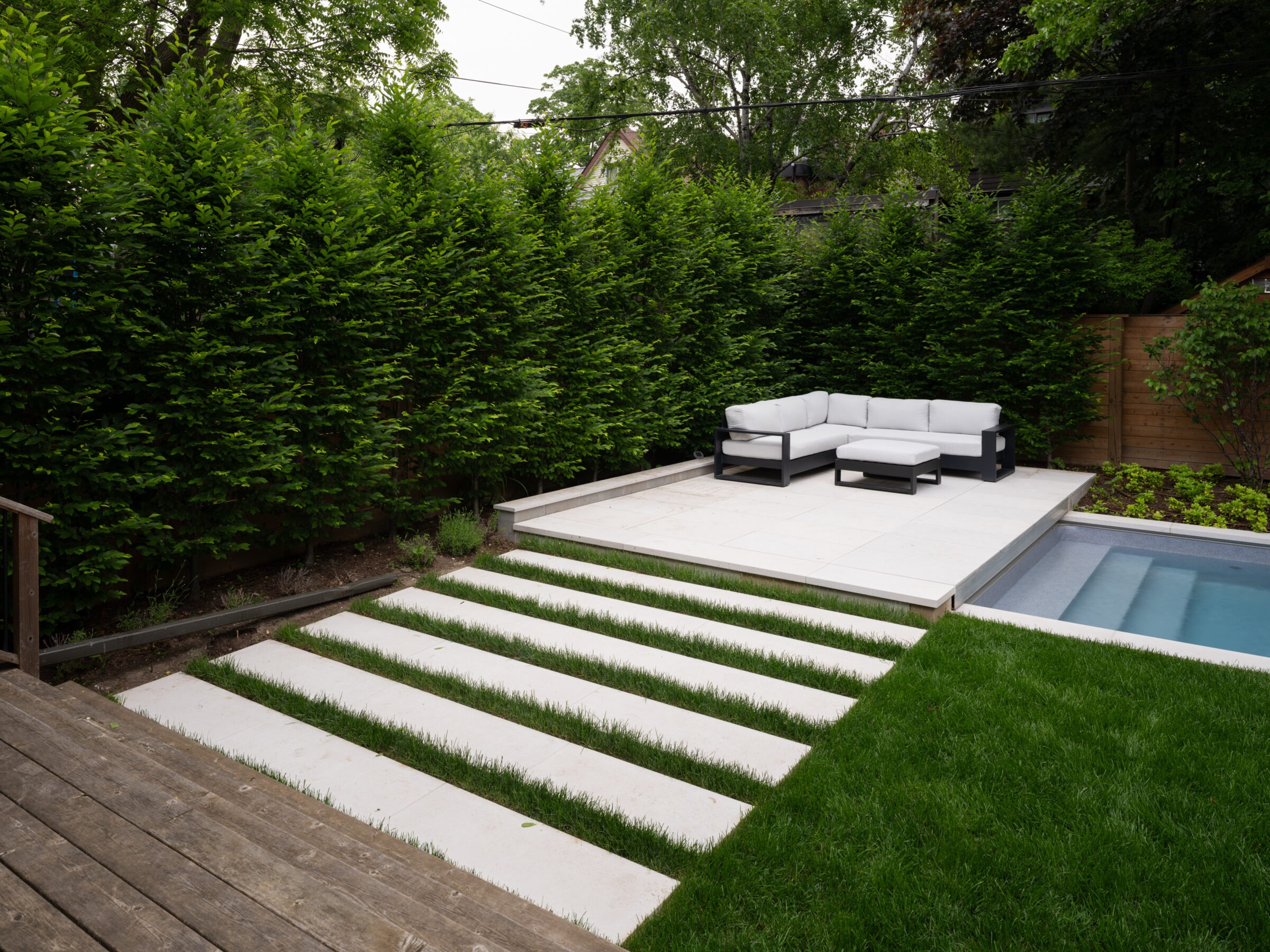 Modern backyard with lush green hedges, white sectional sofa on concrete platform, rectangular pool, and grass alongside white stepping stones. Tranquil setting.