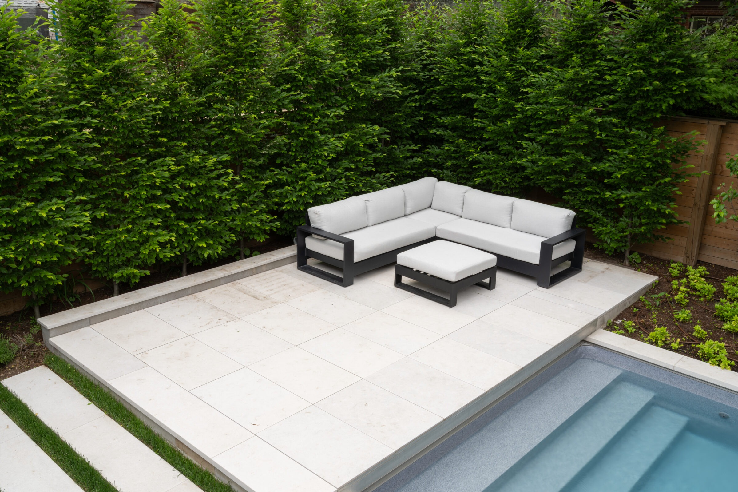 A modern outdoor patio features a white L-shaped sofa and ottoman, adjacent to a swimming pool, surrounded by lush green hedges.