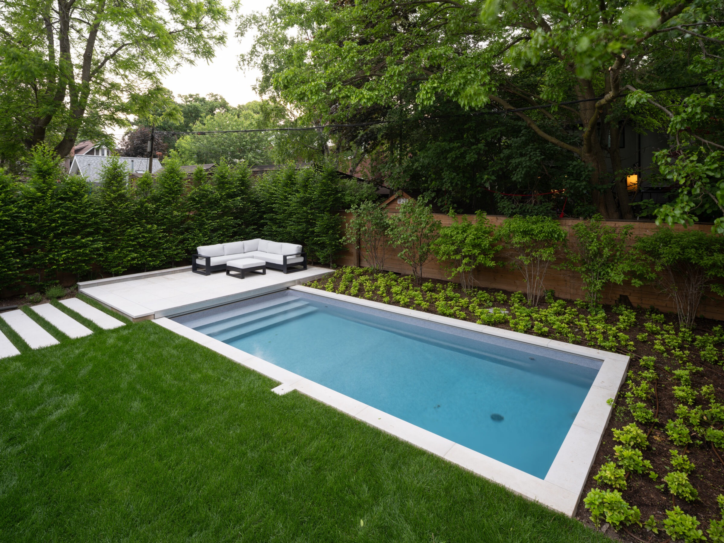 Modern backyard with a rectangular pool, surrounded by lush greenery, trees, and a white outdoor sofa on a tiled patio.