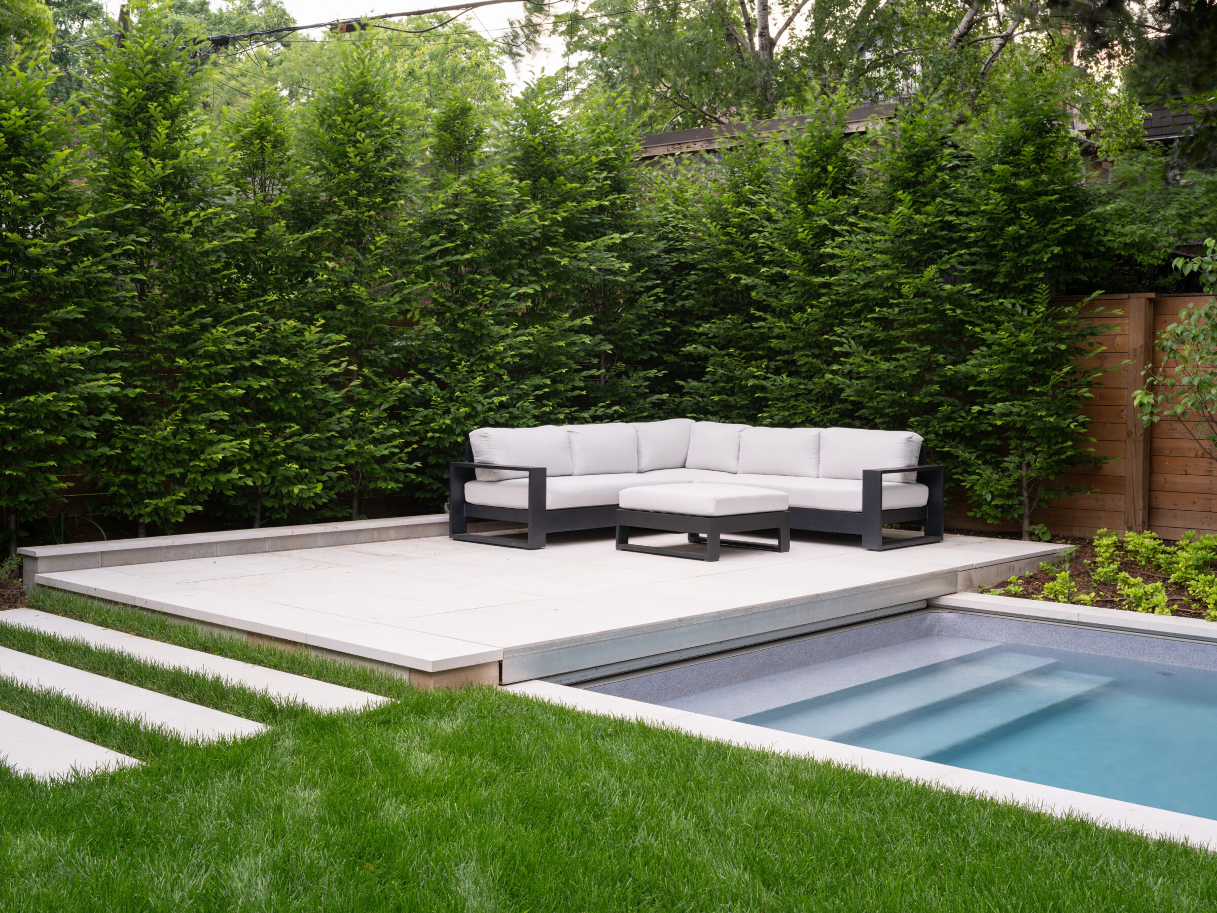 A modern backyard features a white sectional sofa on a raised patio next to a small pool, surrounded by lush greenery and trees.