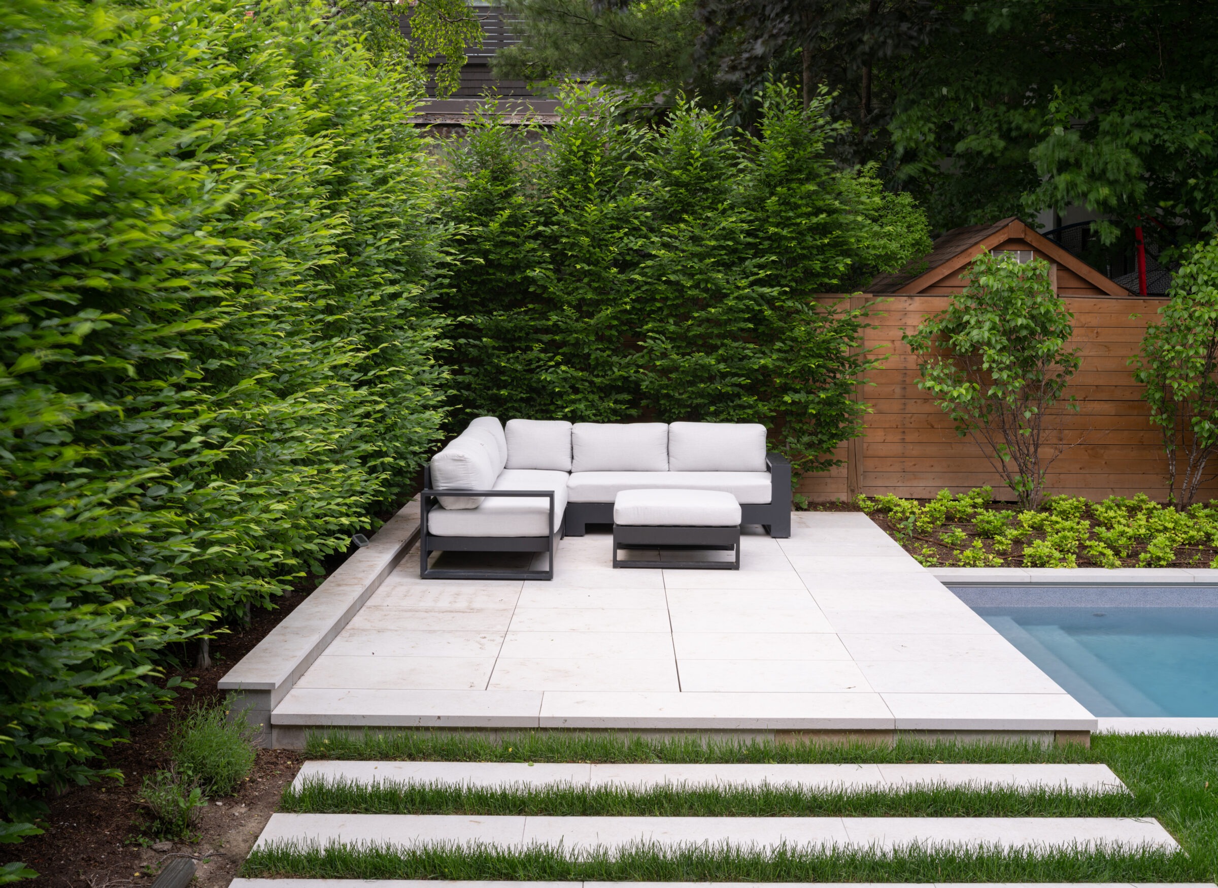 A modern backyard patio with white sectional sofa, surrounded by lush greenery and a small wooden shed, beside a clear blue pool.
