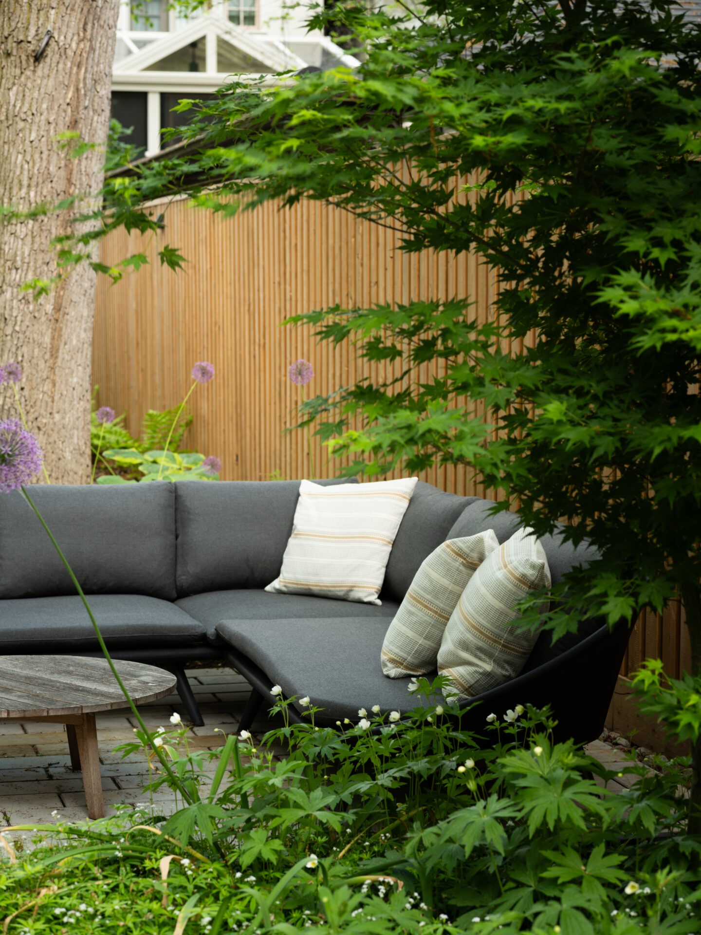A cozy outdoor seating area with gray cushions and striped pillows, surrounded by lush greenery and a wooden fence, creates a tranquil garden atmosphere.